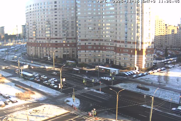 A bustling urban intersection, flanked by large apartment buildings, is visible under bright sunlight, with patches of melting snow and slush on the ground and parked vehicles.