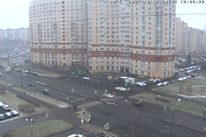 A bustling urban intersection, flanked by large apartment buildings, is visible under bright sunlight, with patches of melting snow and slush on the ground and parked vehicles.