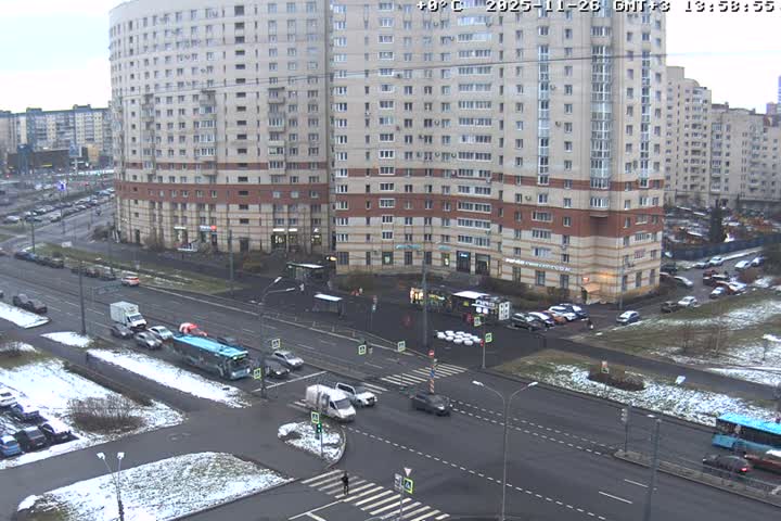 A bustling urban intersection, flanked by large apartment buildings, is visible under bright sunlight, with patches of melting snow and slush on the ground and parked vehicles.