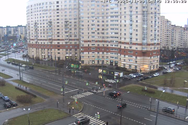 The image shows a bustling urban intersection on an overcast and wet day with temperatures around freezing, featuring multiple cars and pedestrians navigating roads bordered by a large, curved apartment building and other city structures.