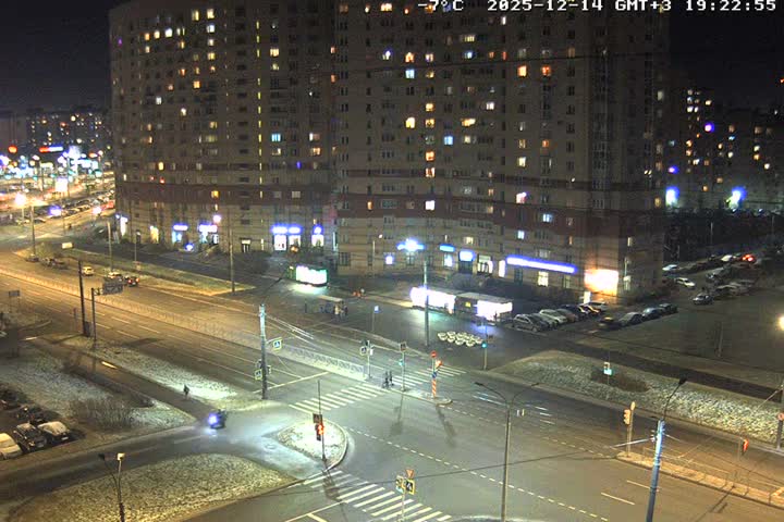 An overhead view captures a busy urban intersection and street lined with multi-story apartment buildings, showing numerous cars, a bus, and pedestrians on a cloudy, damp, and cool day.