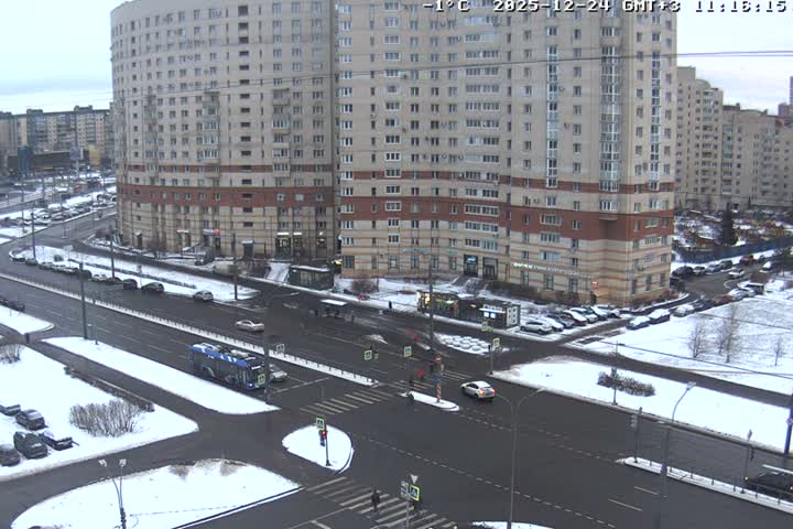 A wide aerial shot captures a snowy urban intersection bustling with vehicles and pedestrians, surrounded by numerous multi-story apartment buildings under an overcast winter sky.