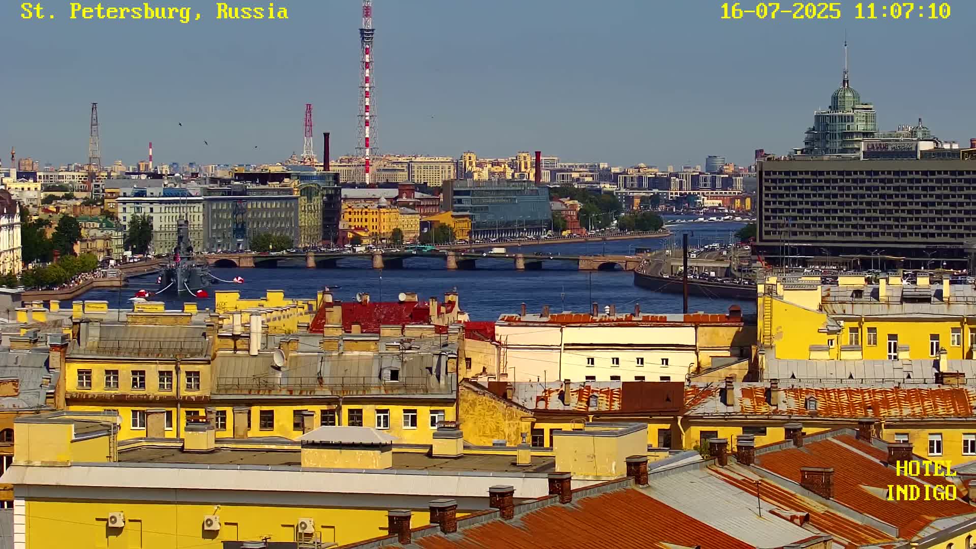 A sunny cityscape shows a river with a bridge and a ship, surrounded by yellow buildings with red-tiled roofs.