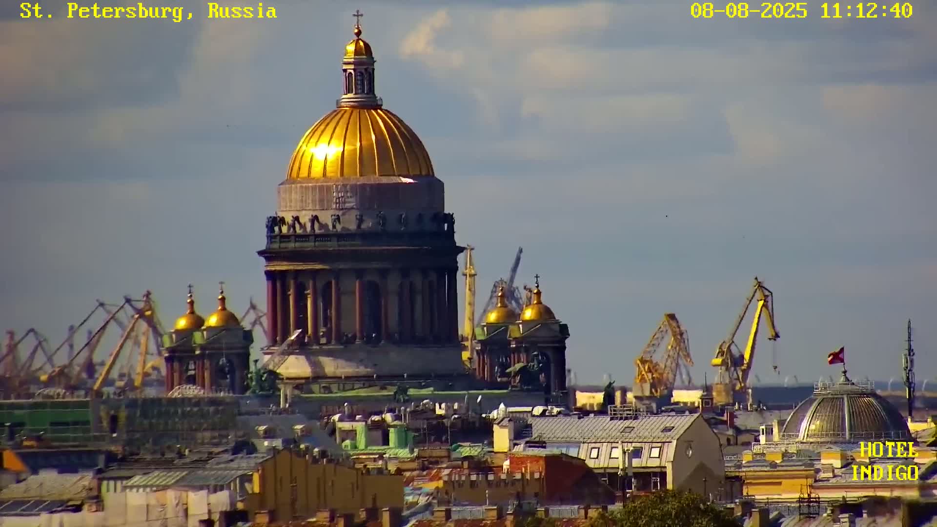A large gold-domed cathedral is seen in the foreground, with a cityscape and harbor cranes visible in the background under a mostly clear sky.