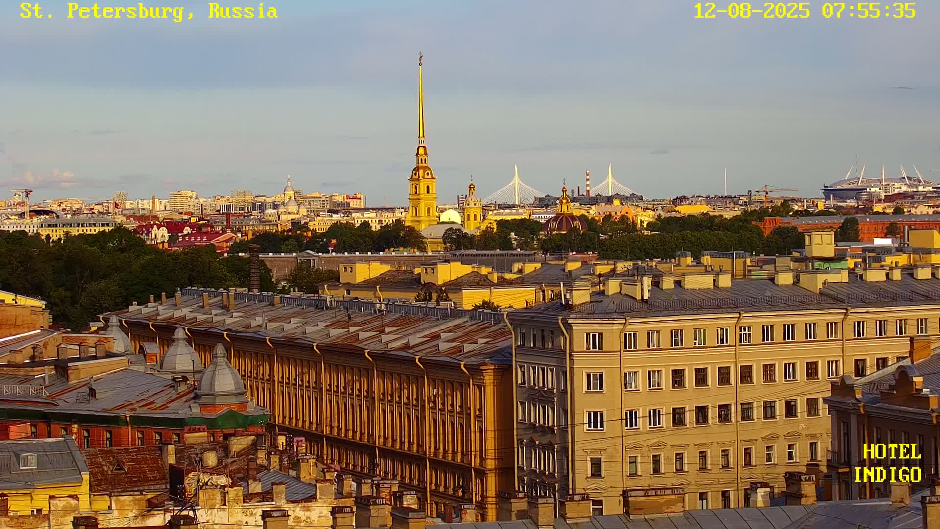 A high-angle view shows a cityscape under a clear, slightly hazy sky, with many yellow buildings in the foreground and a spire-topped church visible in the mid-ground.