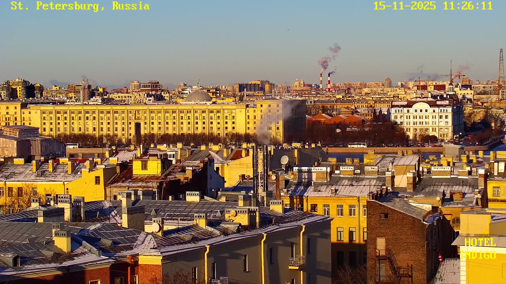 Golden sunlight illuminates a densely packed city on a clear, cold day, with numerous buildings, some featuring snow-dusted roofs, and smoke rising from chimneys into a bright blue sky.