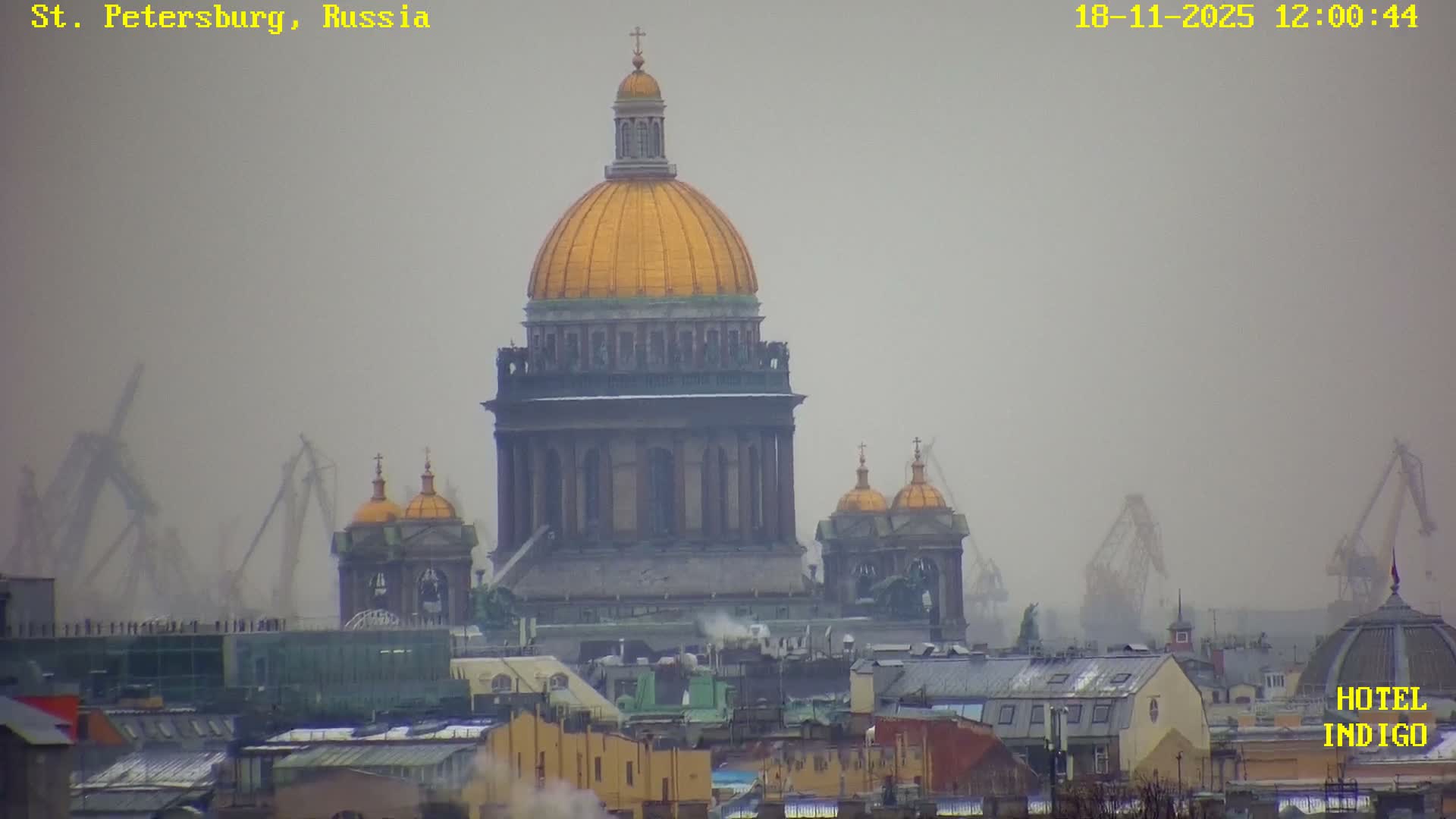 Golden sunlight illuminates a densely packed city on a clear, cold day, with numerous buildings, some featuring snow-dusted roofs, and smoke rising from chimneys into a bright blue sky.