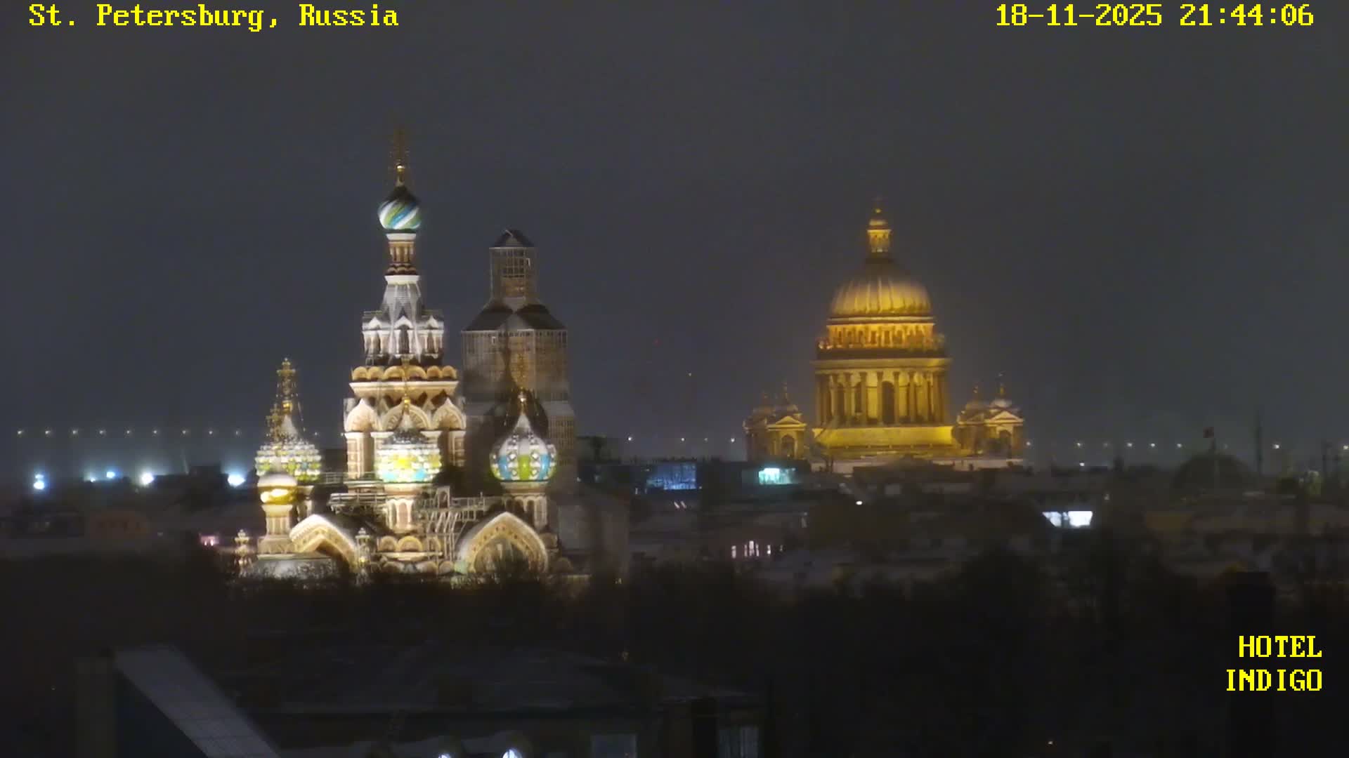 Golden sunlight illuminates a densely packed city on a clear, cold day, with numerous buildings, some featuring snow-dusted roofs, and smoke rising from chimneys into a bright blue sky.