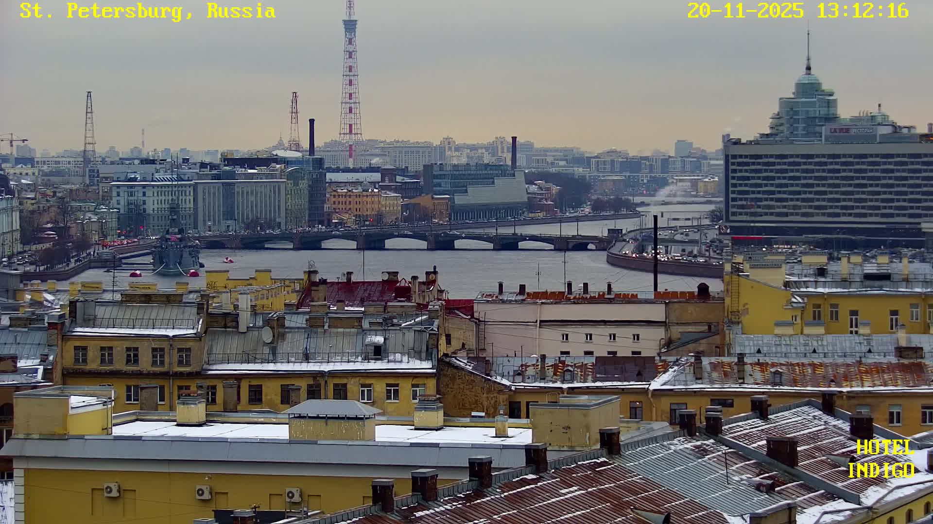 An overcast winter day reveals an urban landscape from above, featuring snow-dusted rooftops in the foreground, a wide river with bridges and a docked ship in the midground, and a distant city skyline with prominent towers under a grey sky.