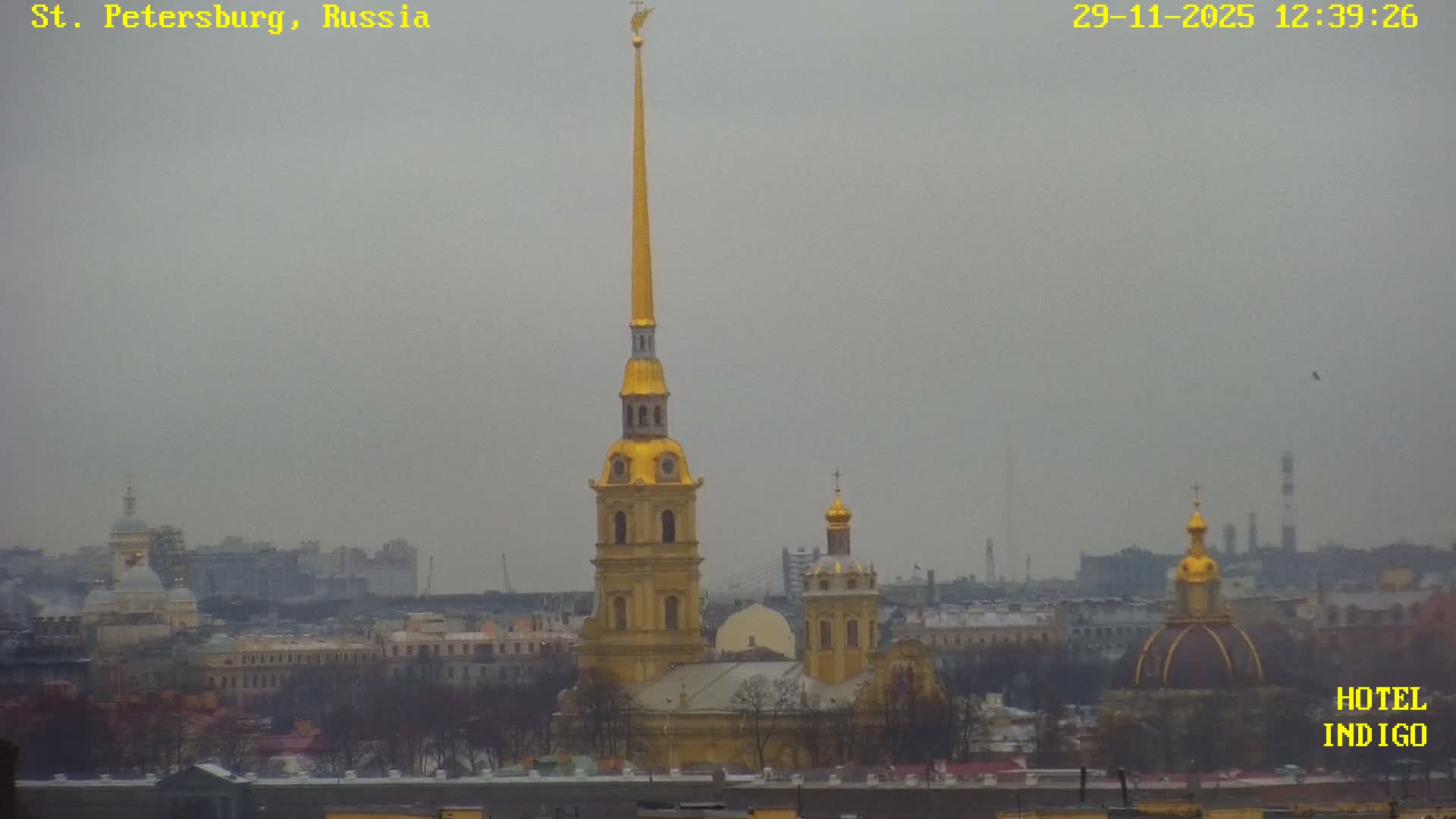 A wide-angle view reveals a city under an overcast and hazy sky with a dusting of snow on rooftops, dominated by the tall golden spire of the Peter and Paul Cathedral and other gilded domes amidst a dense urban landscape of buildings and bare trees.