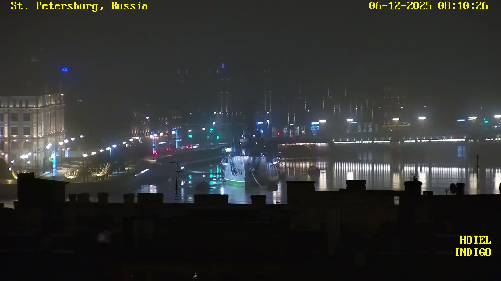A foggy nighttime cityscape depicts illuminated buildings and streets, with numerous lights reflecting on a wide body of water where a large vessel is moored.