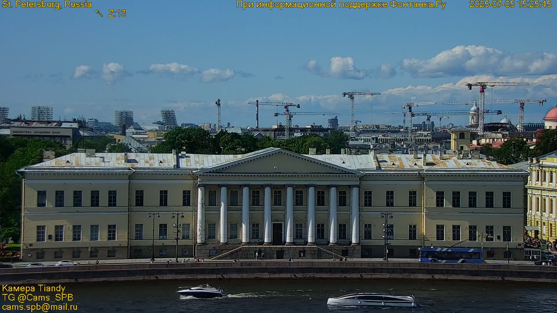 A large, tan building with white columns sits beside a waterway, under a mostly sunny sky with a few clouds and several construction cranes visible in the distance.
