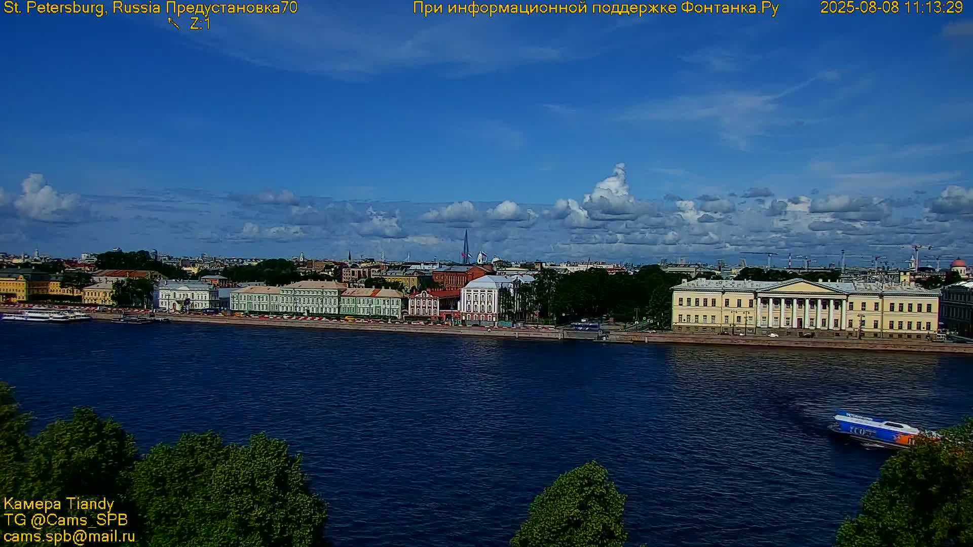 A partly cloudy day shows a city waterfront with several buildings along a river, and a hydrofoil is visible in the water.