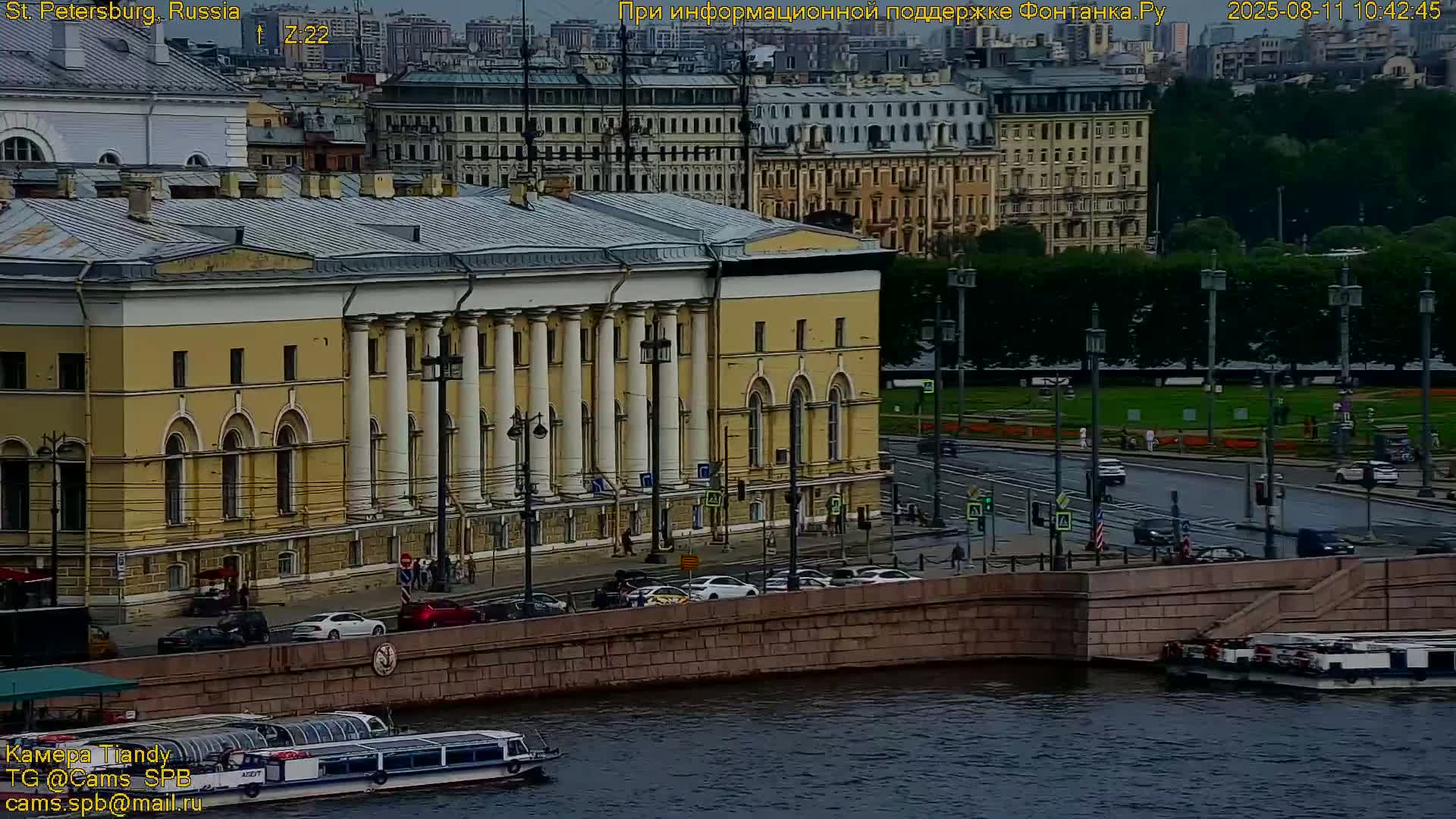 A large, tan building with columns stands along a waterway, where boats are docked and cars drive on a nearby bridge under an overcast sky.
