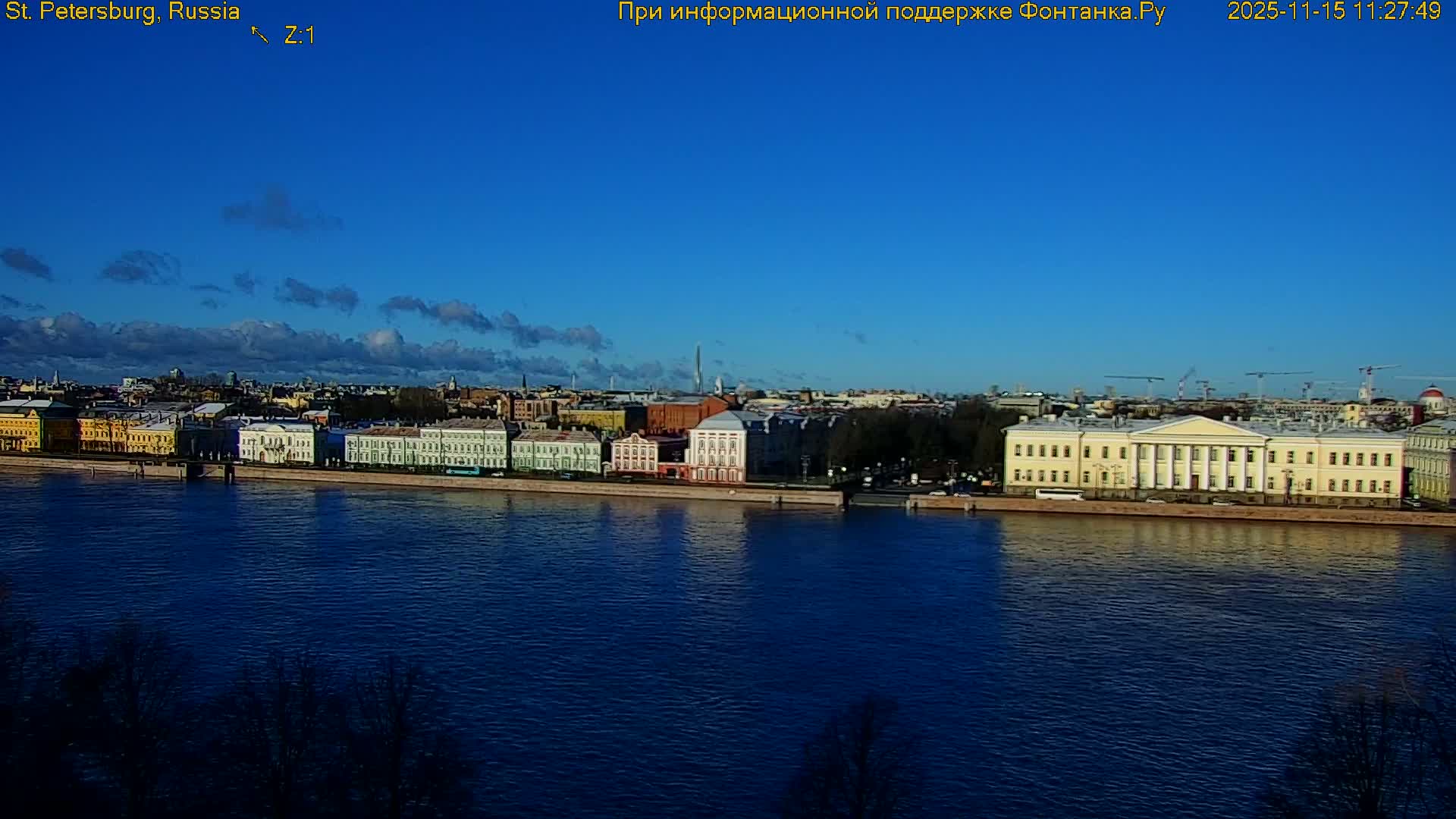 The image shows a city riverfront on a sunny day, featuring a wide blue river reflecting a clear sky with scattered white clouds, bordered by a long stretch of historic buildings and a distant cityscape.