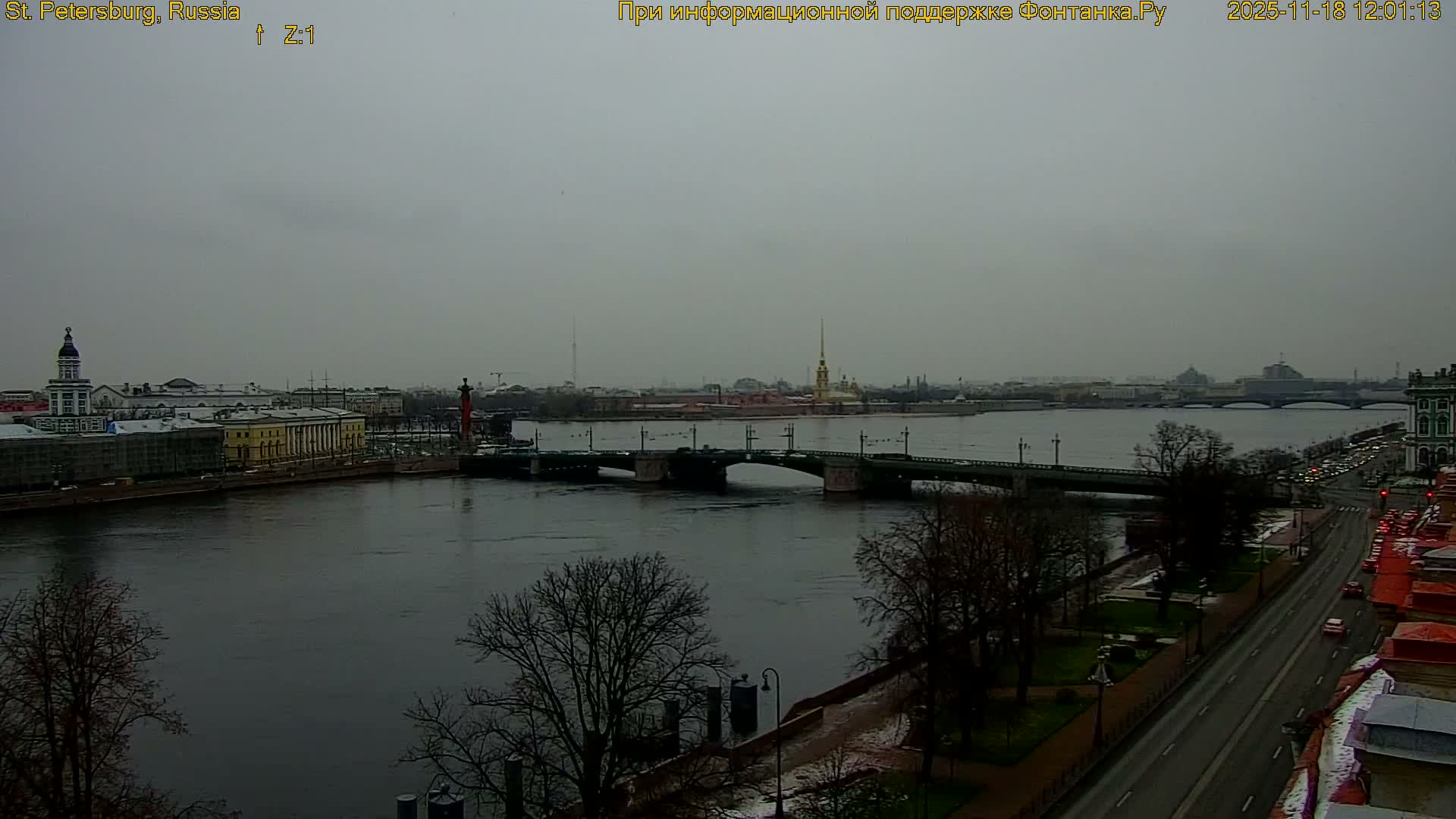 The image shows a city riverfront on a sunny day, featuring a wide blue river reflecting a clear sky with scattered white clouds, bordered by a long stretch of historic buildings and a distant cityscape.