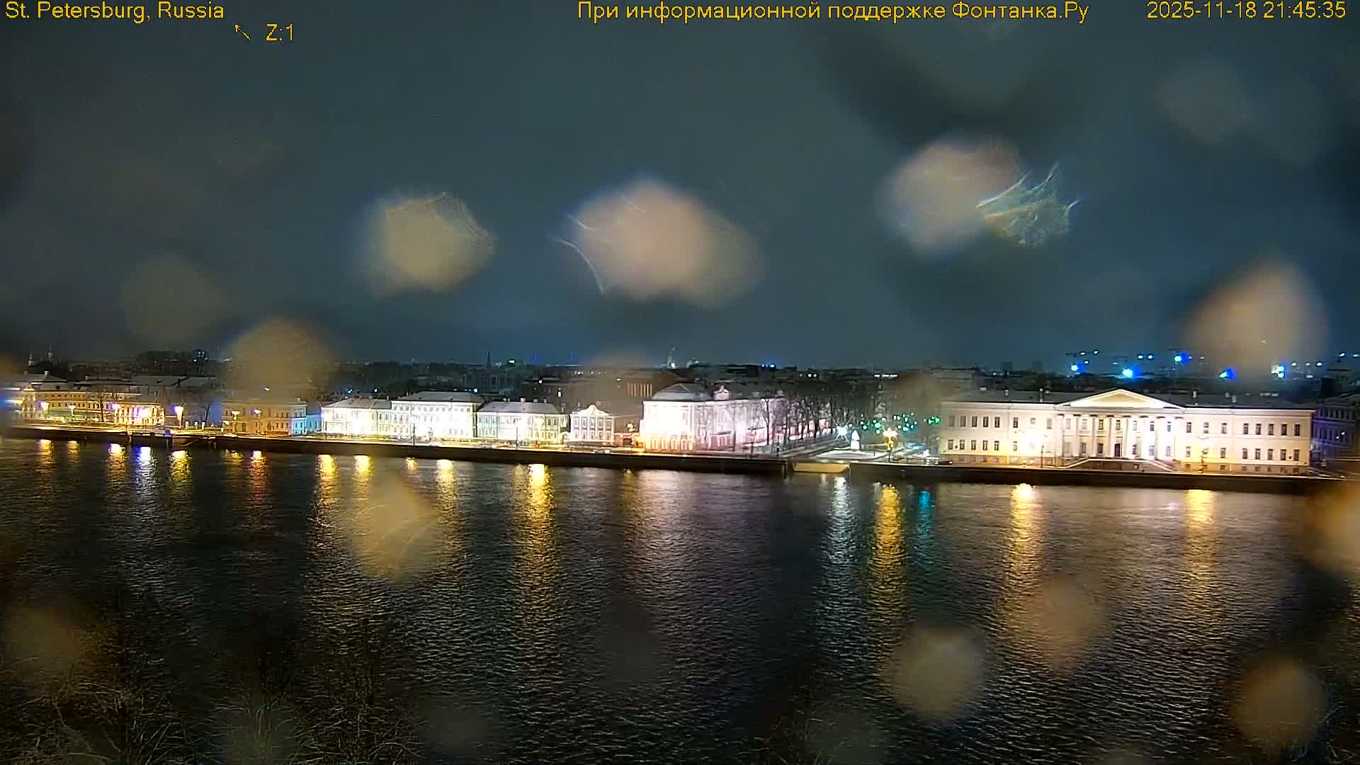 The image shows a city riverfront on a sunny day, featuring a wide blue river reflecting a clear sky with scattered white clouds, bordered by a long stretch of historic buildings and a distant cityscape.