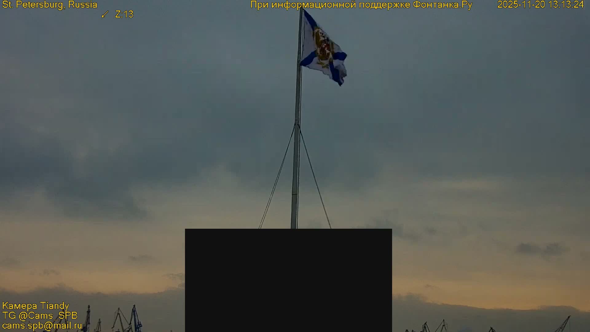 A white, blue, and yellow flag flies on a pole against a predominantly overcast sky with hints of twilight colors, with port cranes just visible on the horizon.