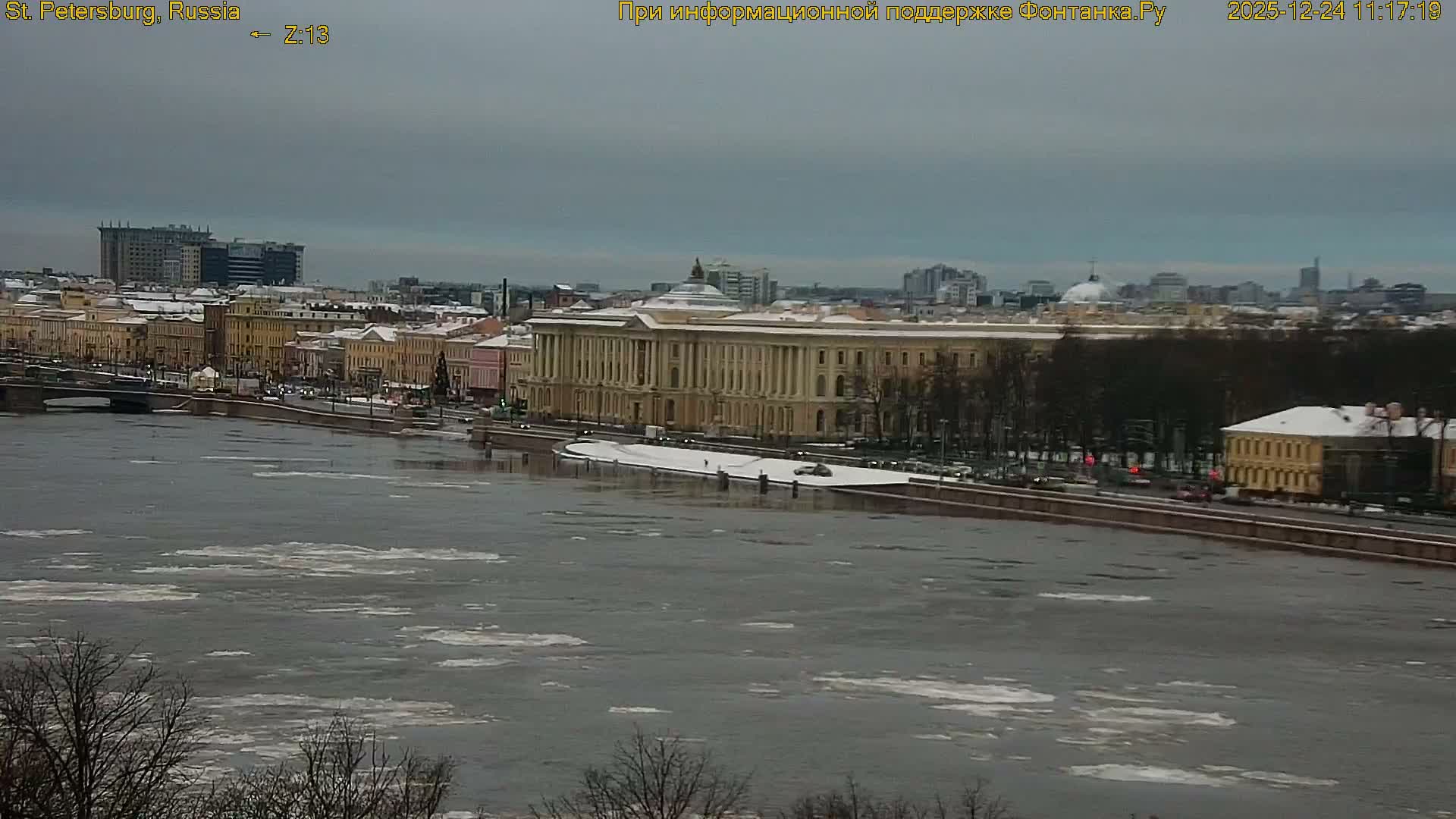 A wide, partially frozen river with ice floes flows past a snow-dusted cityscape featuring historic buildings, a bridge, and bare trees under a grey, overcast winter sky.