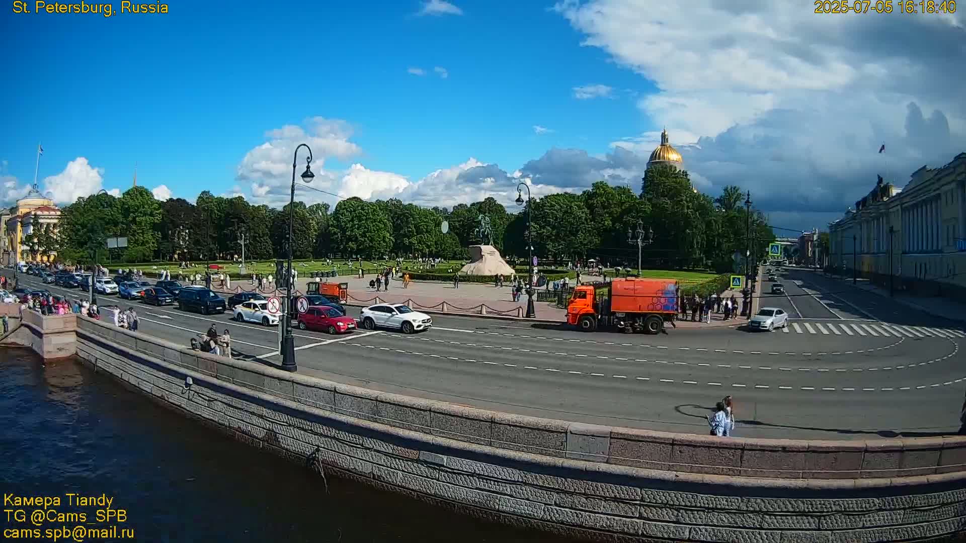 A sunny day with partly cloudy skies shows a city street scene with traffic, pedestrians, and a large park with a monument in the background.