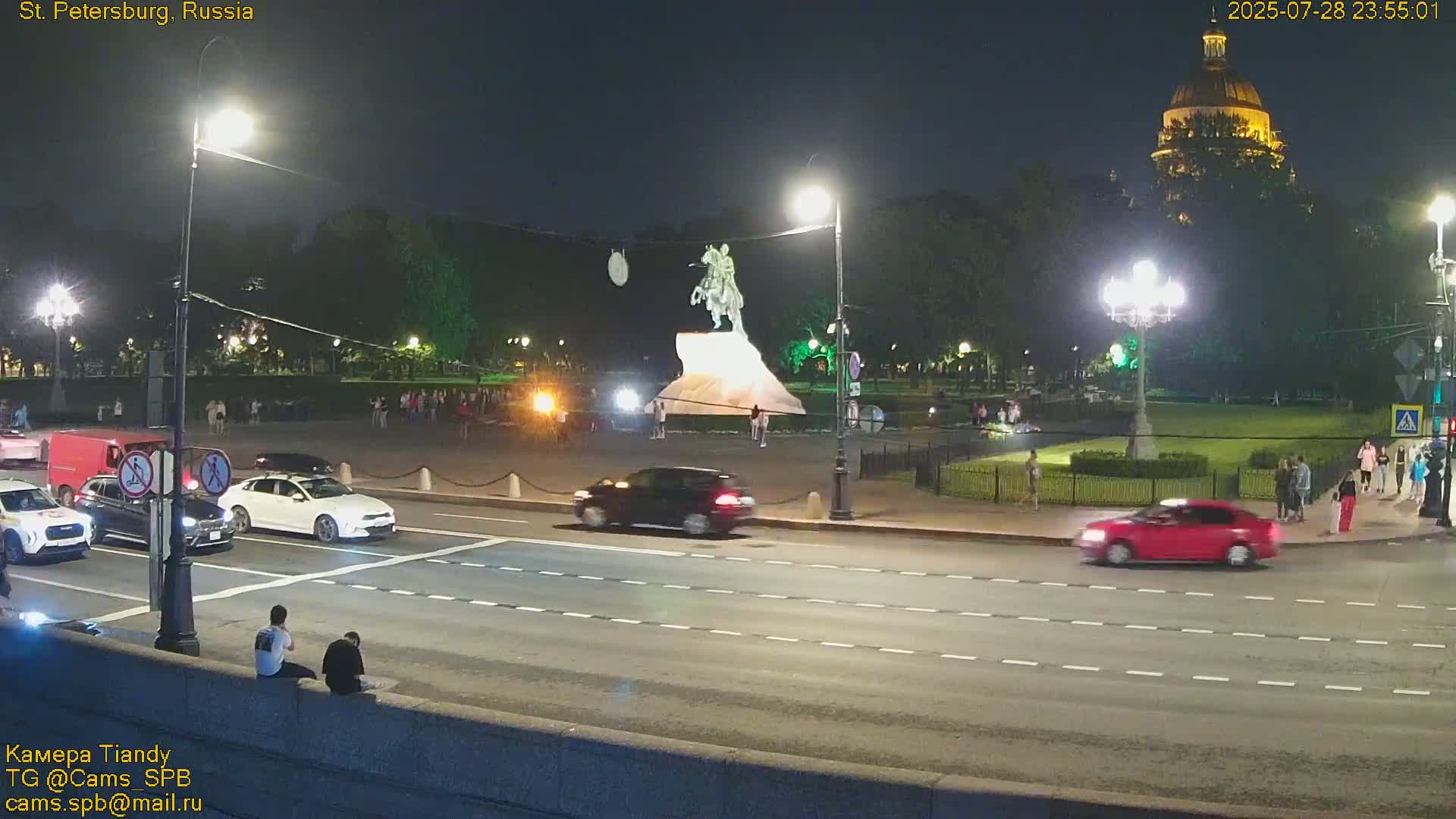 At night, several cars drive on a multi-lane road past a statue on a pedestal in a plaza with people walking around, all illuminated by streetlights, with a large domed building in the background.