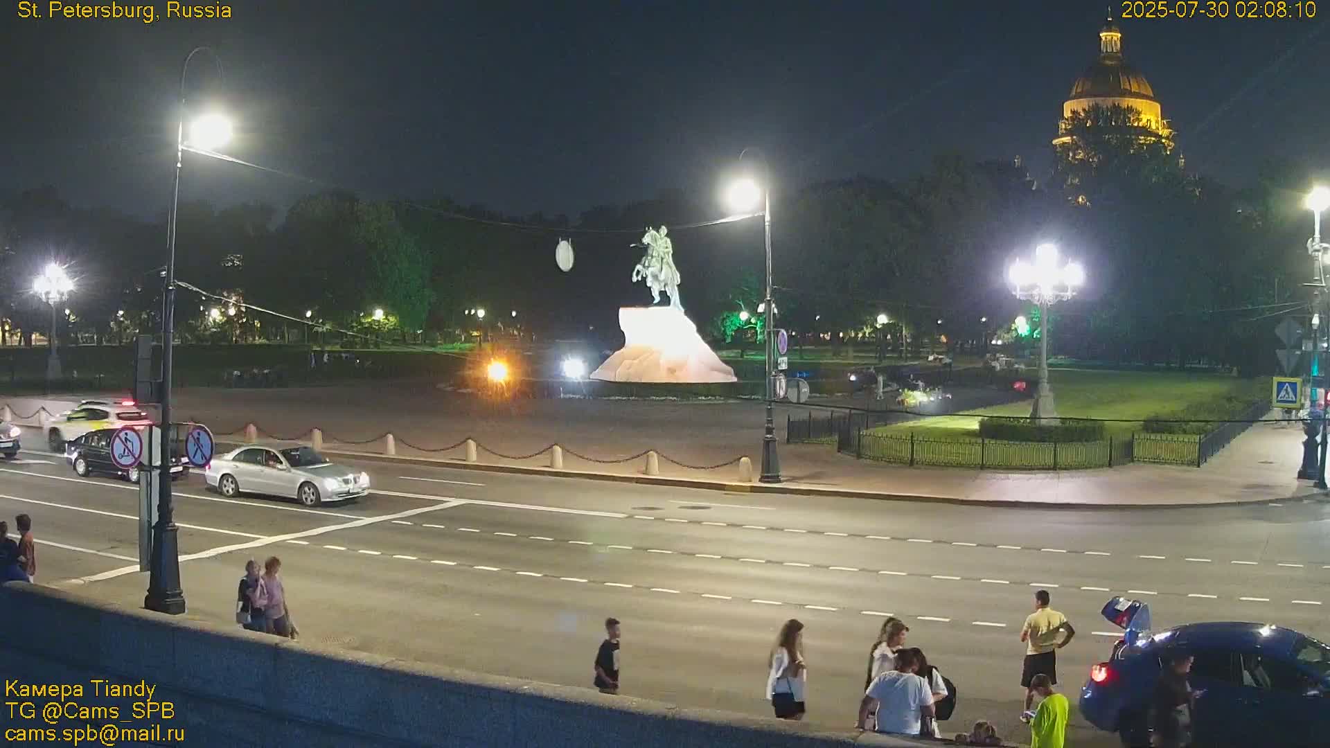 At night, several cars drive on a multi-lane road past a statue on a pedestal in a plaza with people walking around, all illuminated by streetlights, with a large domed building in the background.