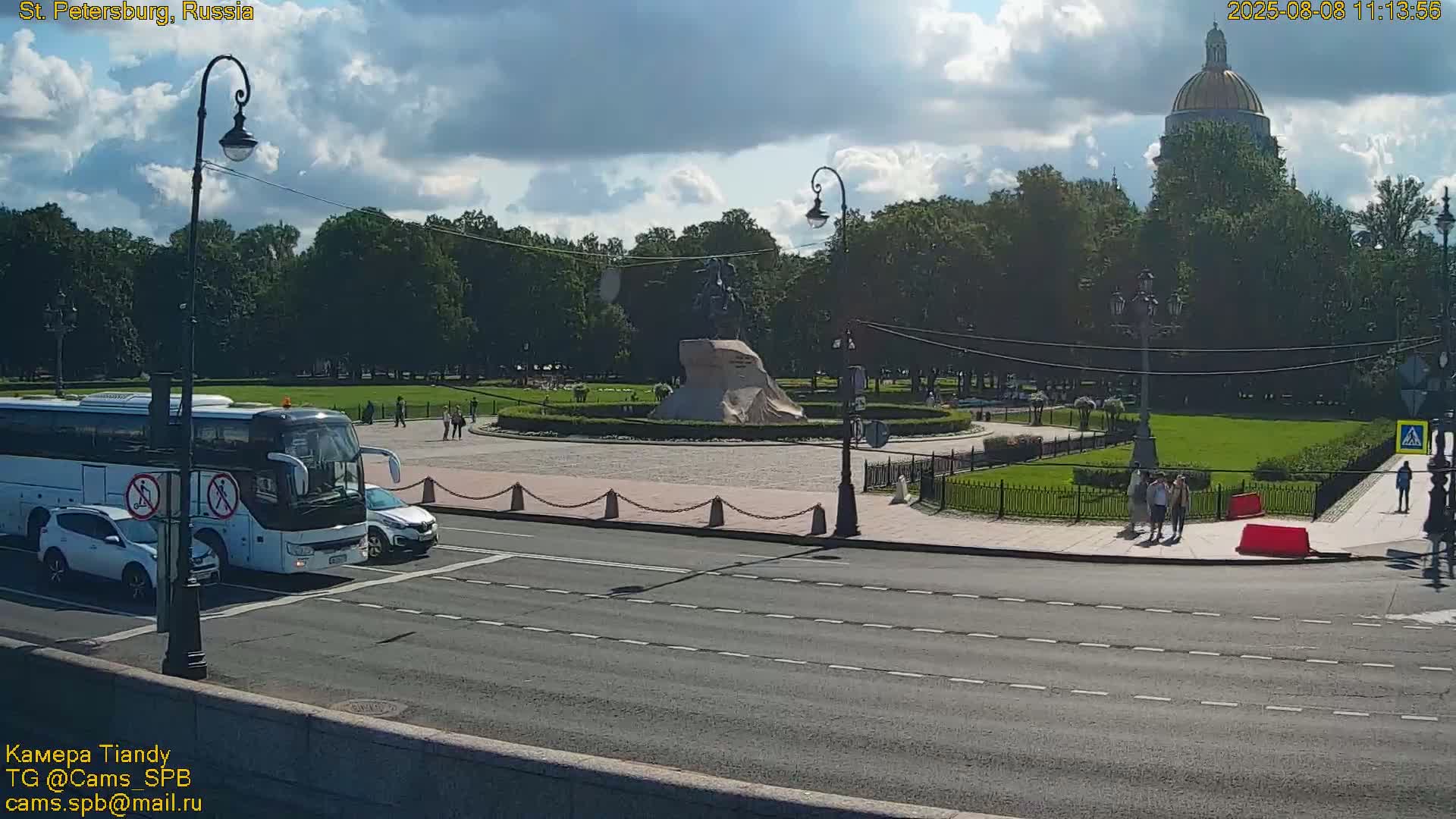 A partly cloudy day shows a city street scene with a large bus and several cars, a monument covered in a cloth, a large green park, and a domed building in the background.