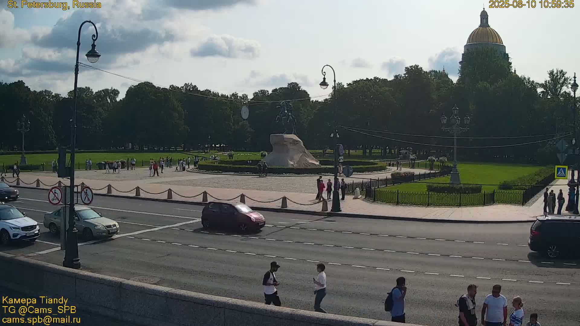 A partly cloudy day shows a city square with a large equestrian statue,  surrounding green space, a building with a golden dome in the background, and cars and pedestrians on a road.