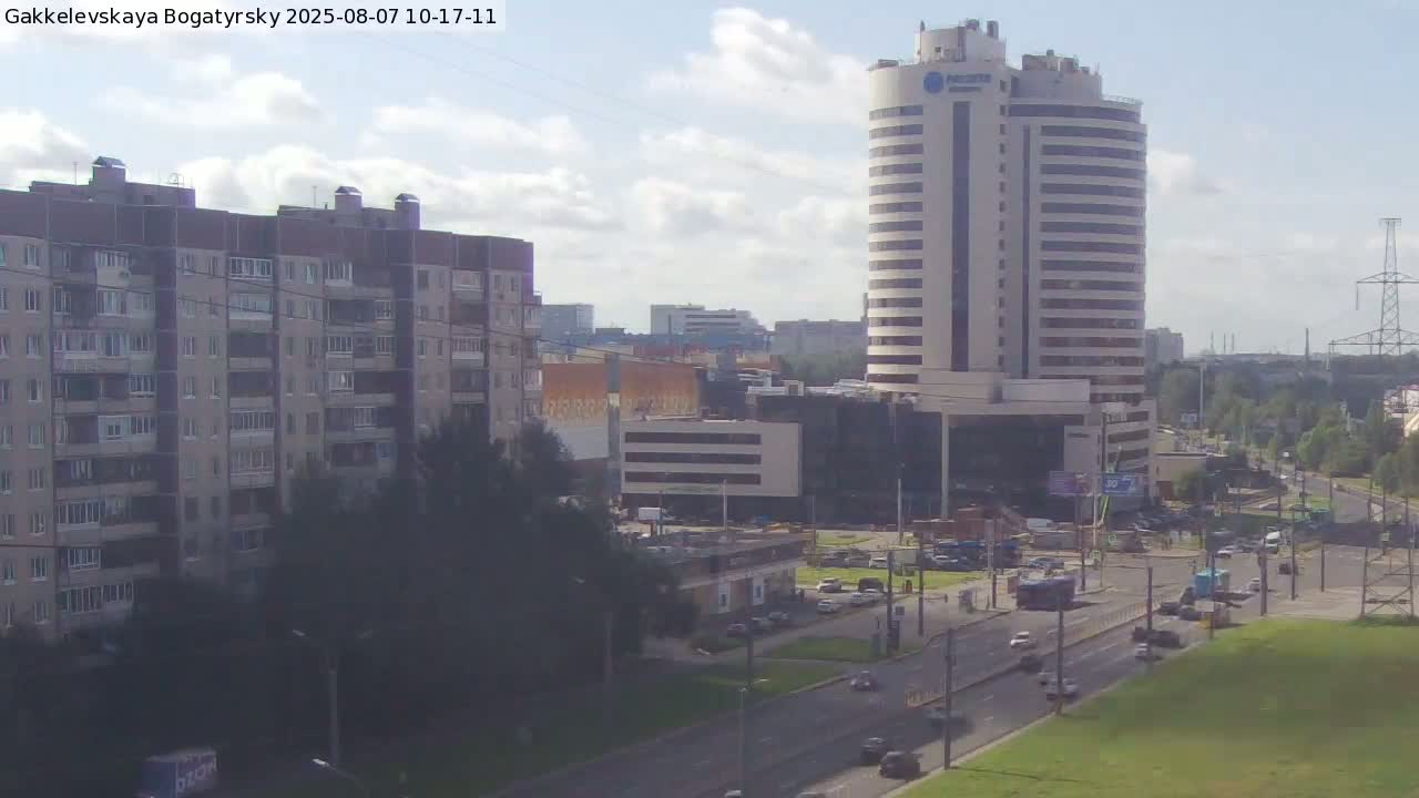 A partly cloudy day reveals a cityscape with multi-story apartment buildings, a tall cylindrical office building, and a busy road with cars and a bus.