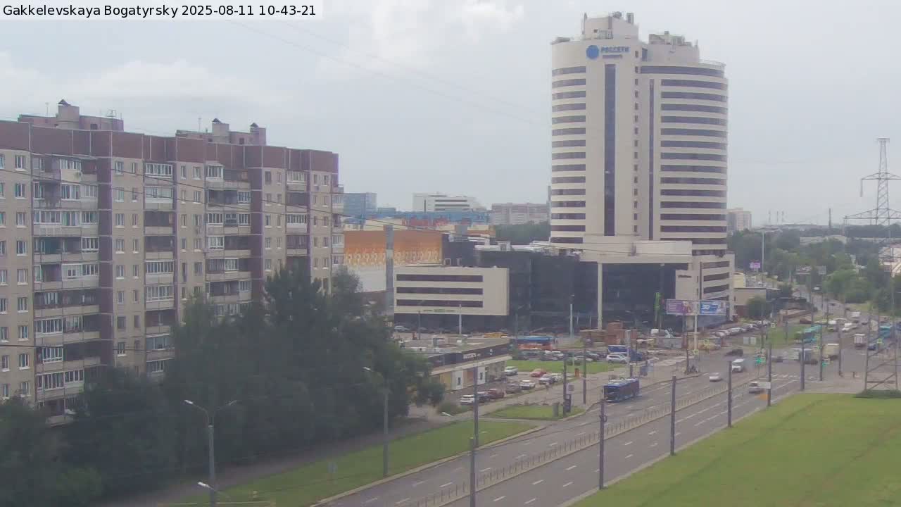 A city street scene on an overcast day shows a tall, light-colored building next to a multi-story apartment building, with a roadway separating them and containing various vehicles.