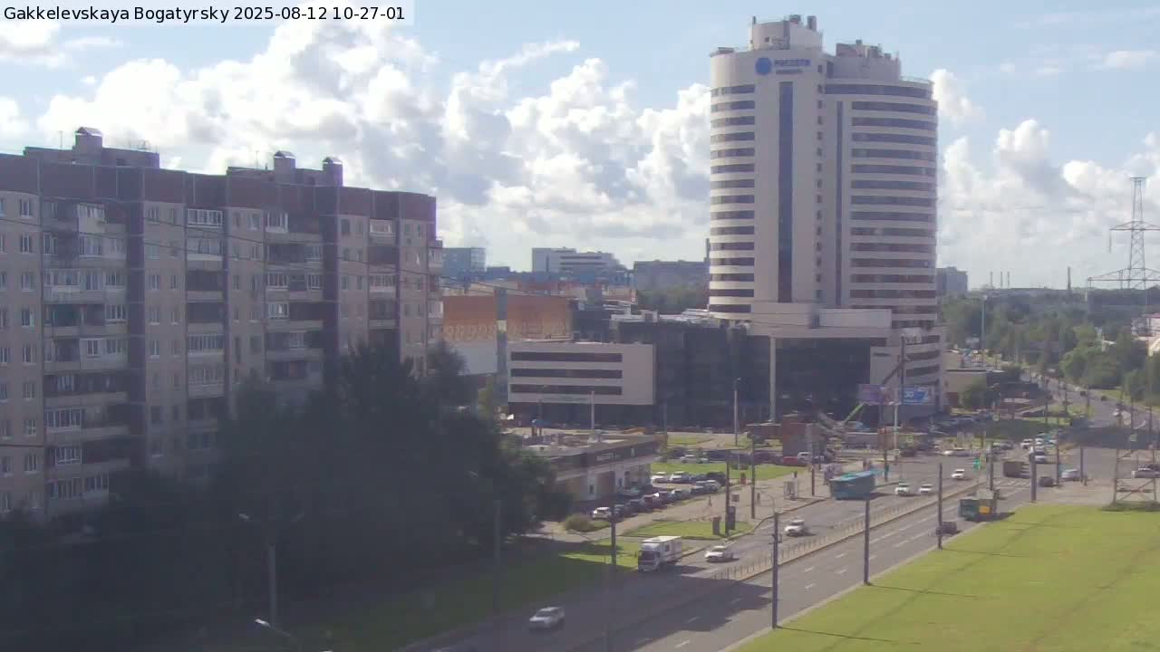 A partly cloudy day reveals a cityscape featuring multi-story apartment buildings, a tall, curved office building, and a busy road with several cars and a bus.