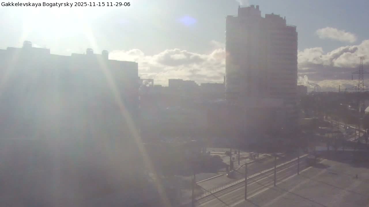 A bright, hazy sun casts lens flare over a sprawling cityscape, revealing tall buildings, a multi-lane road, and distant industrial structures under a partly cloudy sky.