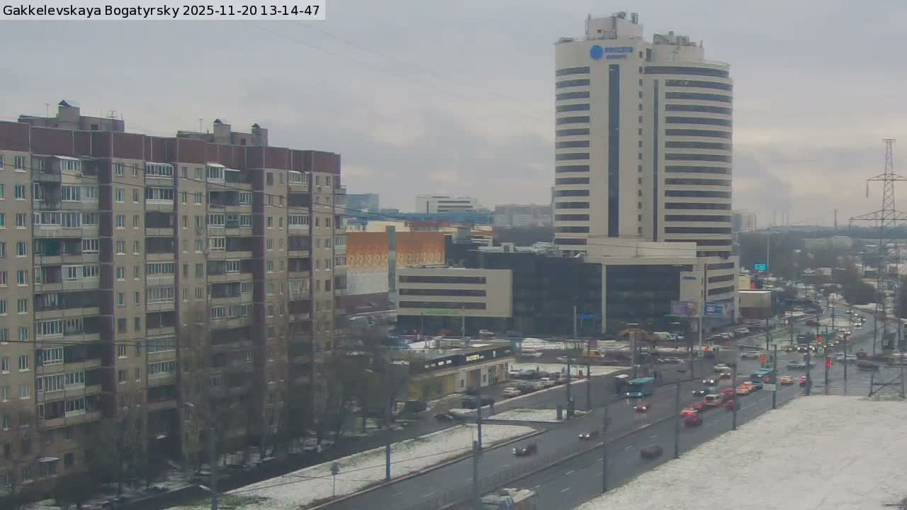 An urban scene depicts a busy multi-lane street with traffic, flanked by a large apartment complex on the left and a tall modern high-rise building on the right, all under a grey, overcast sky with a light dusting of snow visible on the ground.