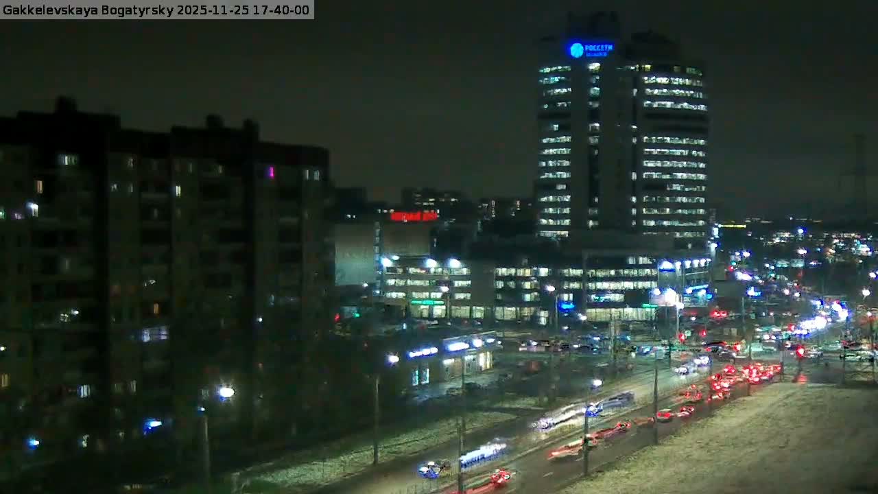 An urban scene depicts a busy multi-lane street with traffic, flanked by a large apartment complex on the left and a tall modern high-rise building on the right, all under a grey, overcast sky with a light dusting of snow visible on the ground.