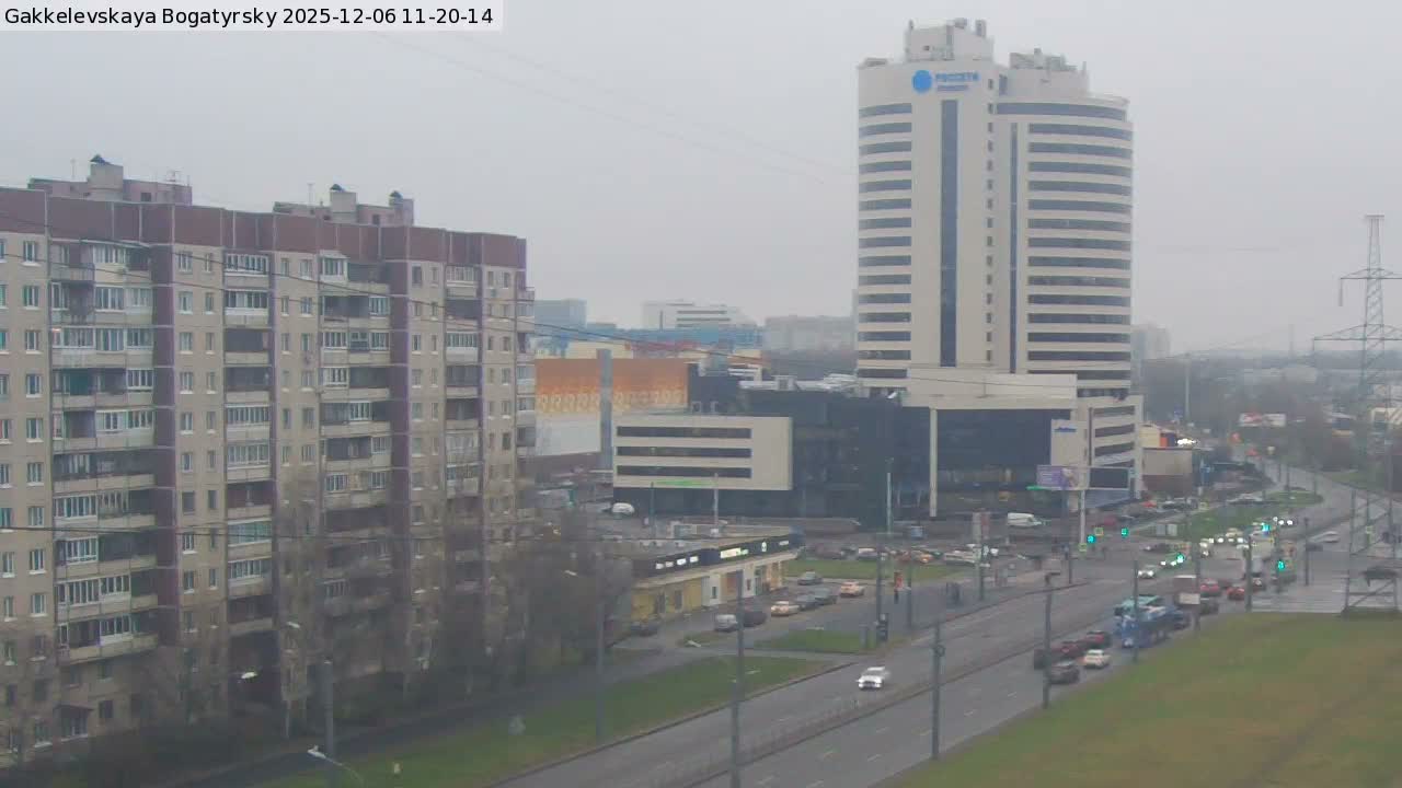 An overcast and damp city scene features a long apartment building on the left, a tall modern high-rise on the right, and a wide multi-lane road with moderate traffic in the foreground.
