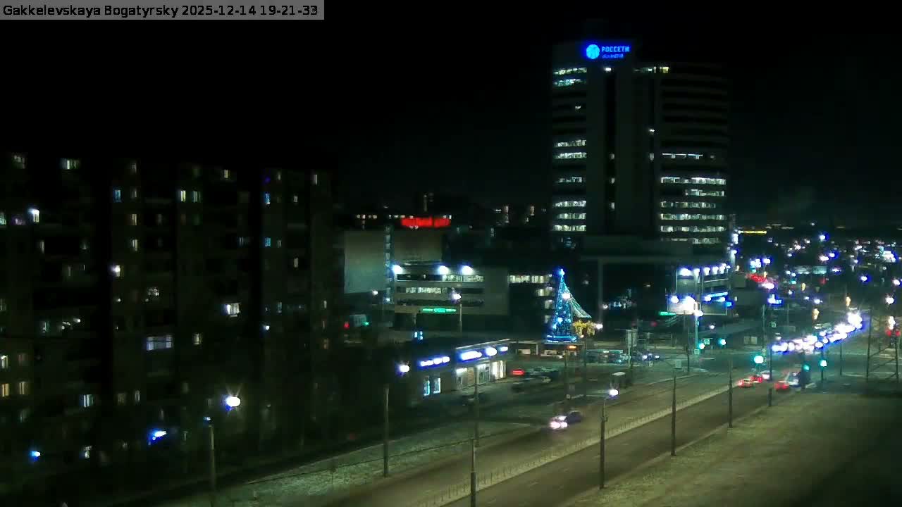 An overcast and damp city scene features a long apartment building on the left, a tall modern high-rise on the right, and a wide multi-lane road with moderate traffic in the foreground.