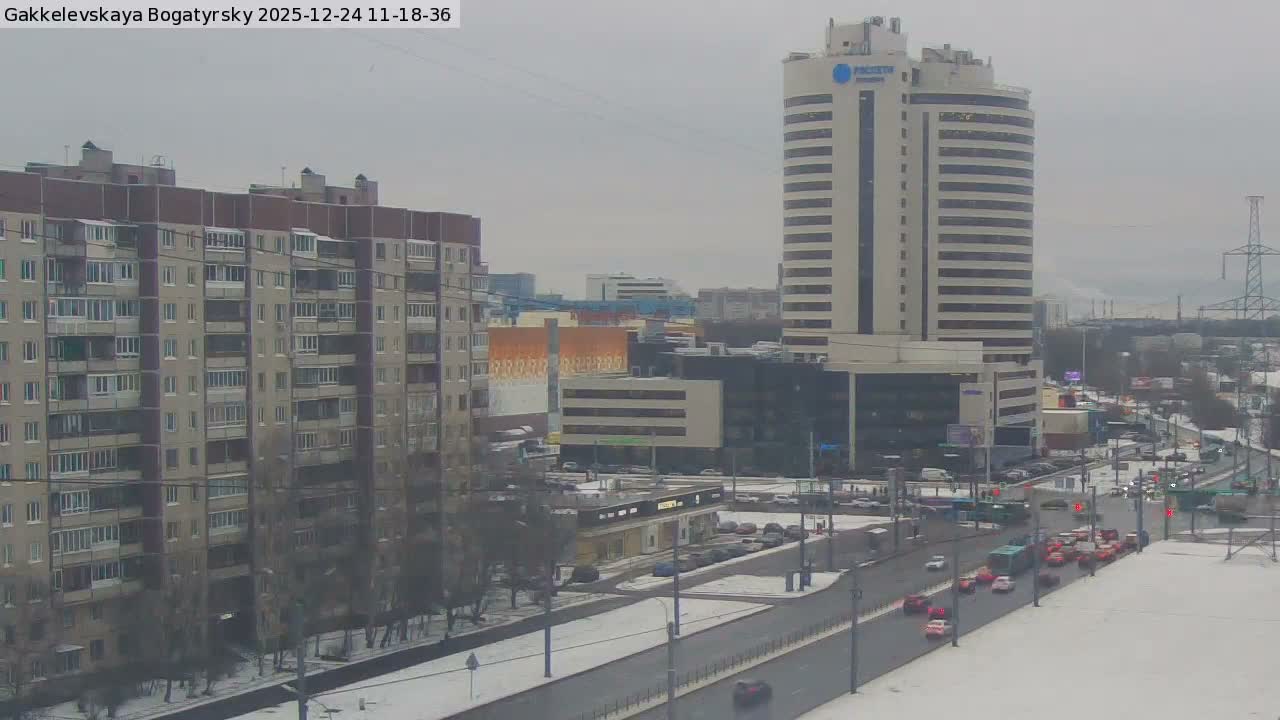The image displays a snowy urban landscape on a cloudy winter day, featuring a multi-lane road with traffic in the foreground, flanked by a large residential building on the left and a prominent high-rise office building on the right.