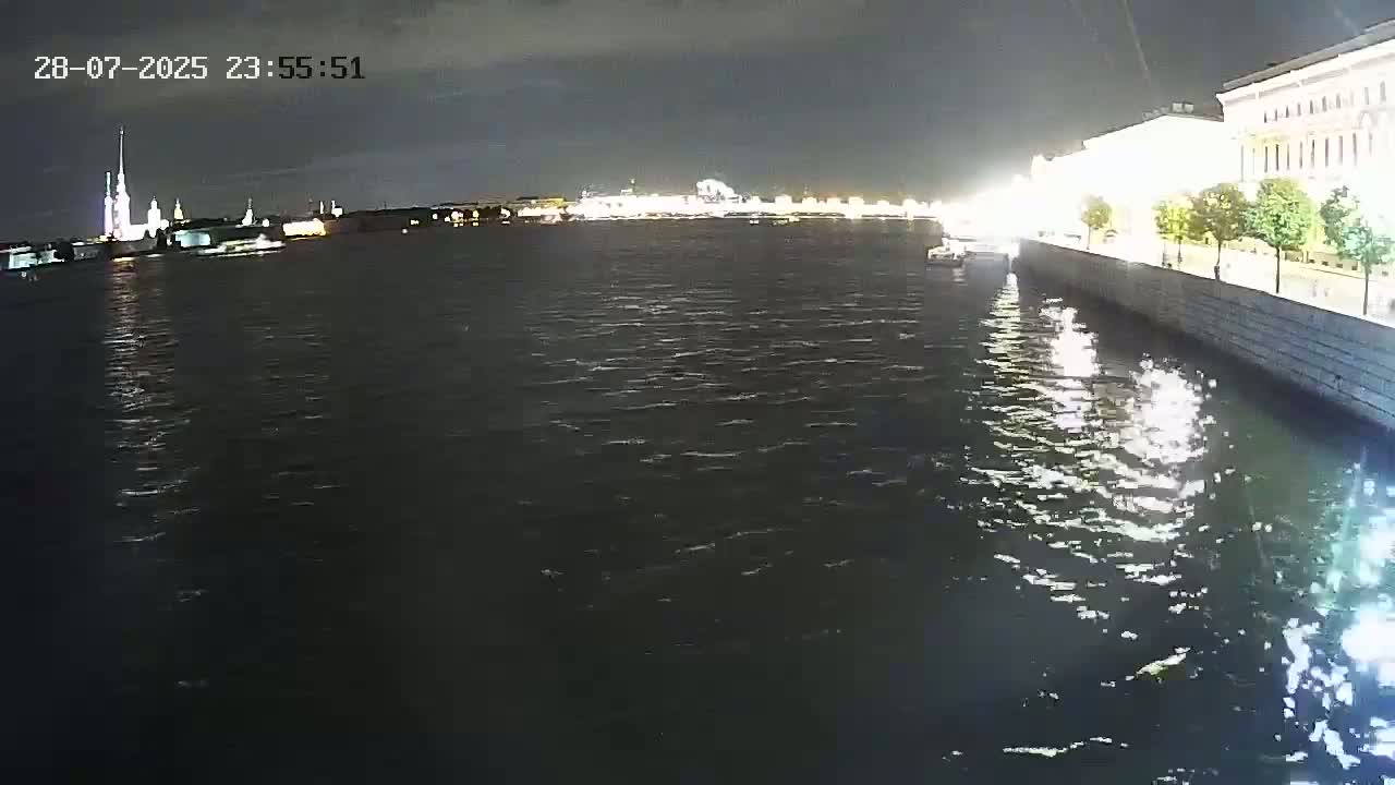 A nighttime view of a dark body of water with city lights reflecting on its surface, showing a cityscape with buildings along the waterfront under a dark sky.