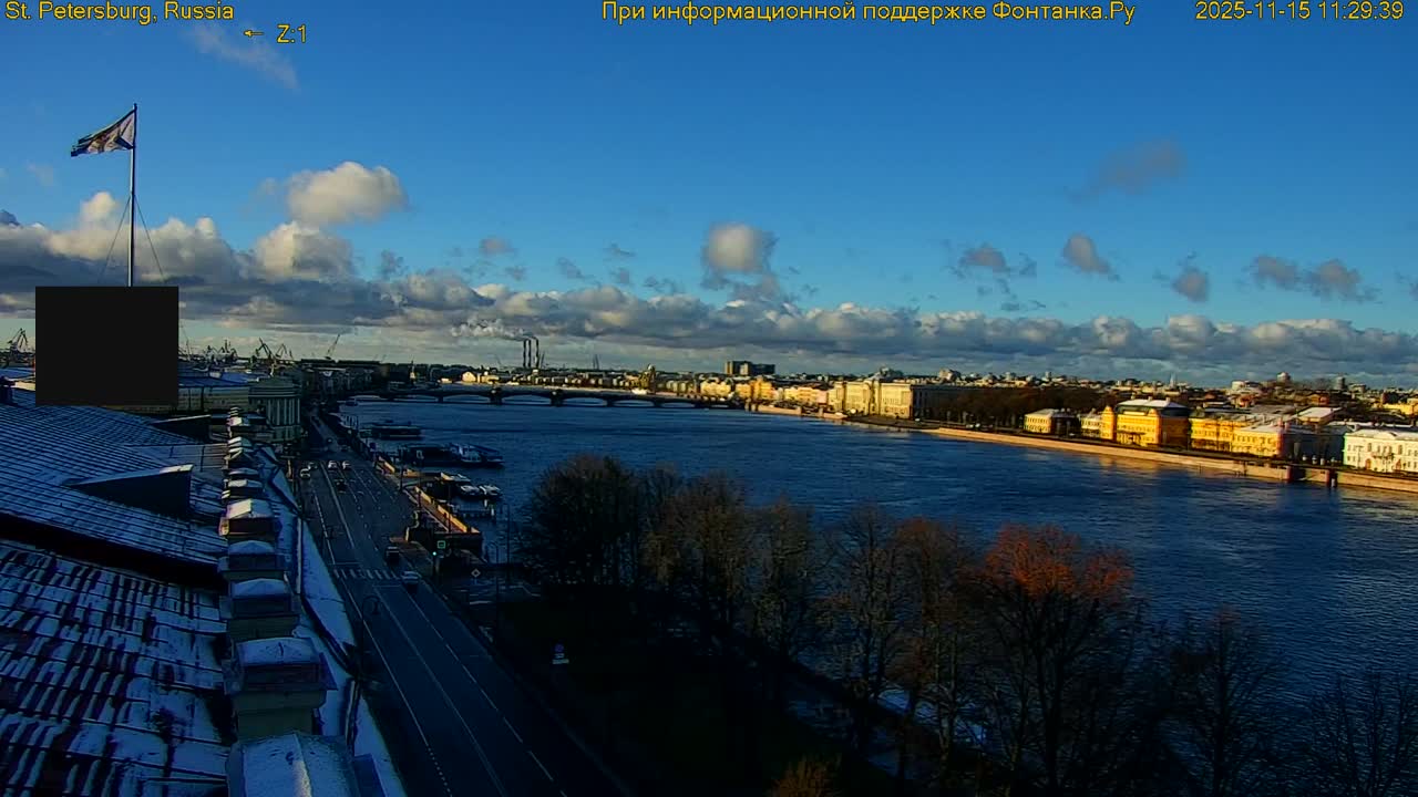 A wide river flows through a city with buildings lining its banks and a road alongside, featuring bare trees and rooftops dusted with what appears to be light snow or frost, all bathed in sunlight under a partly cloudy blue sky, suggesting crisp, clear weather.