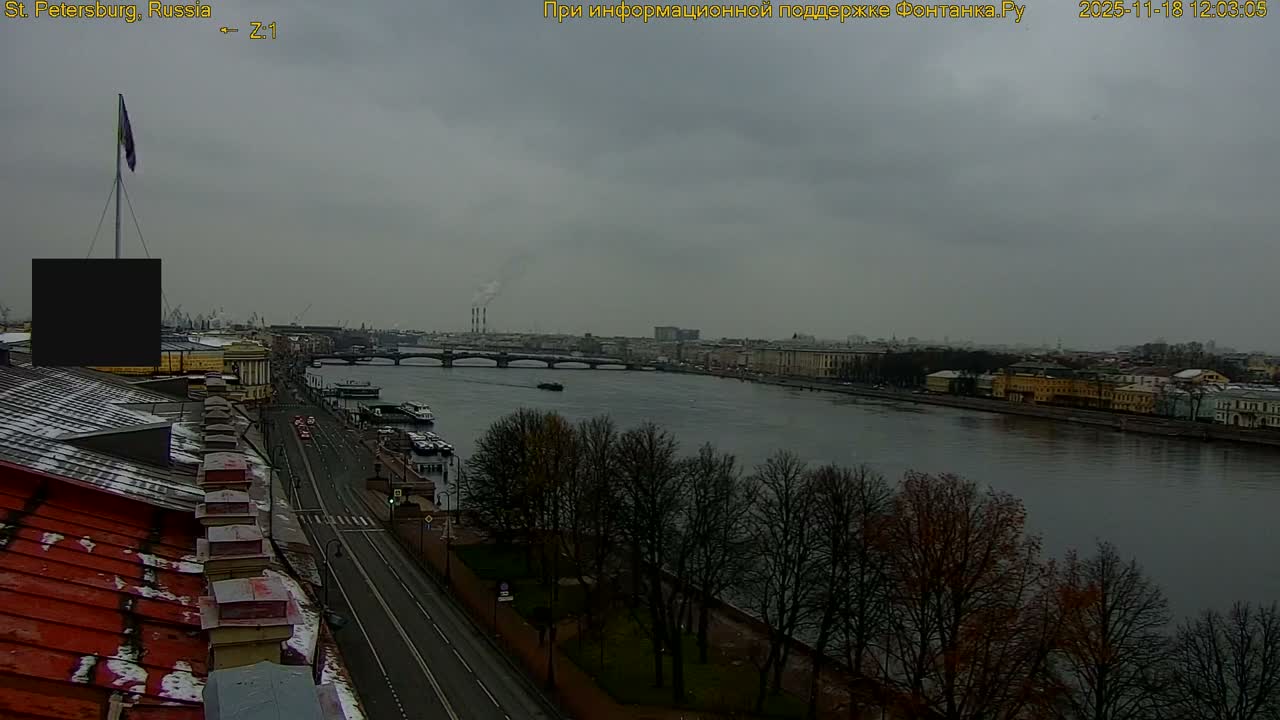 A wide river flows through a city with buildings lining its banks and a road alongside, featuring bare trees and rooftops dusted with what appears to be light snow or frost, all bathed in sunlight under a partly cloudy blue sky, suggesting crisp, clear weather.