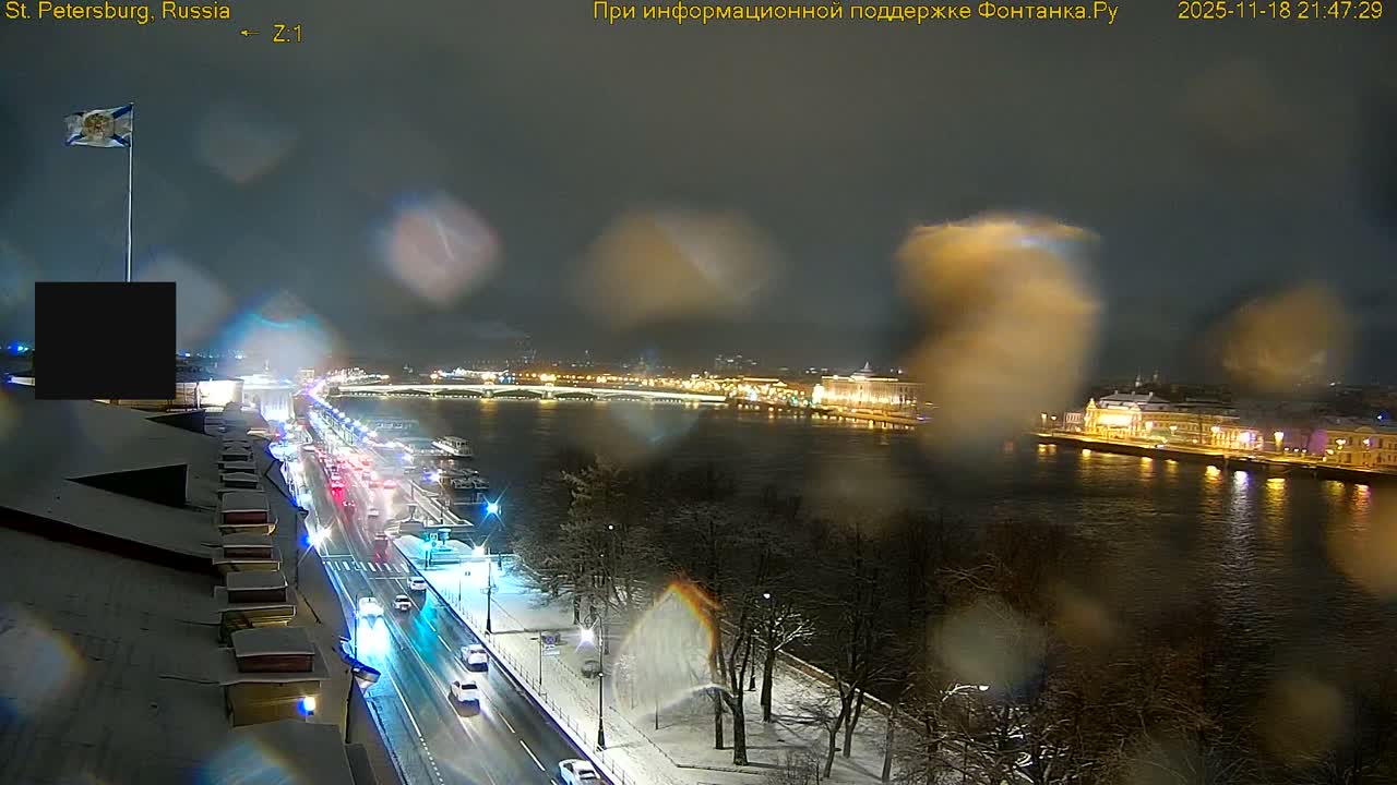 A wide river flows through a city with buildings lining its banks and a road alongside, featuring bare trees and rooftops dusted with what appears to be light snow or frost, all bathed in sunlight under a partly cloudy blue sky, suggesting crisp, clear weather.
