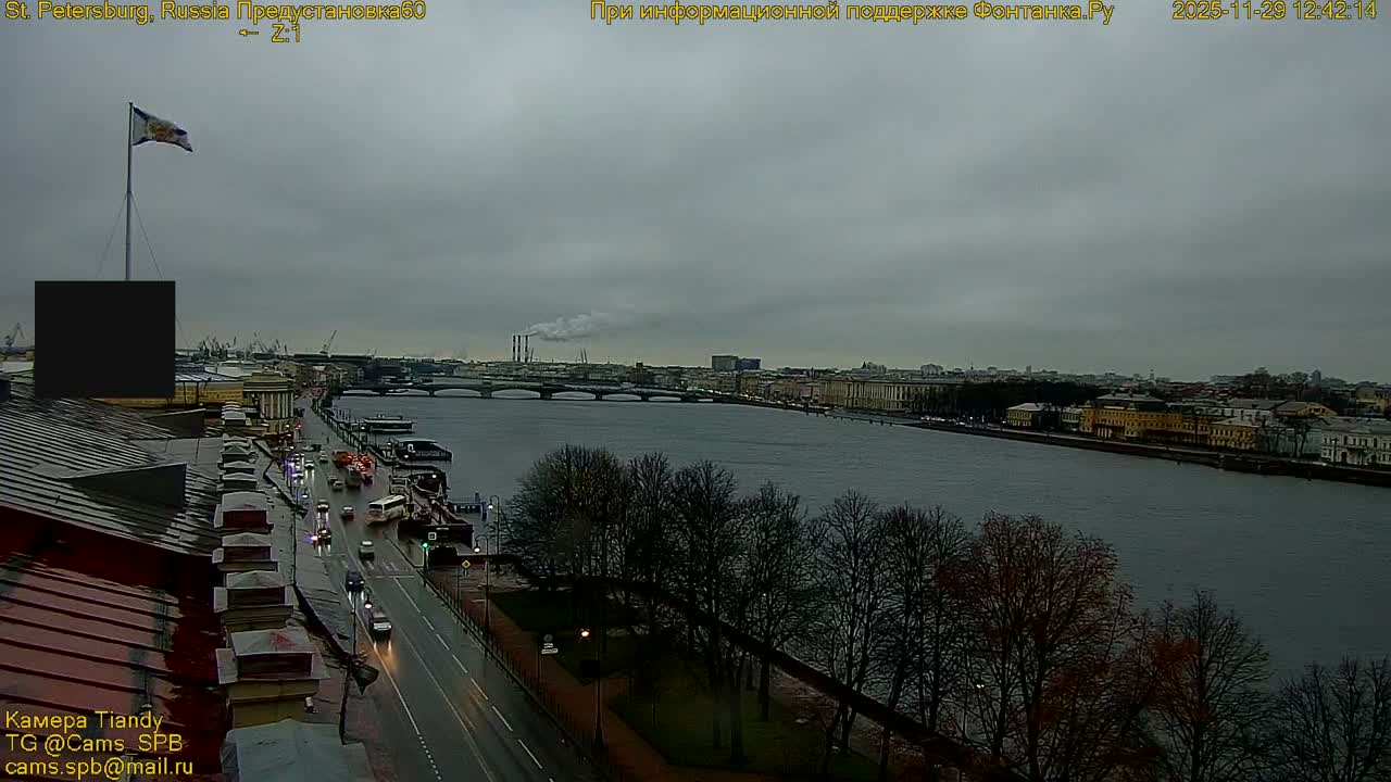 A panoramic view over a wide city river, with traffic moving along a wet road on the left, bridges spanning the water, and a cityscape stretching into the distance under an overcast and damp sky.