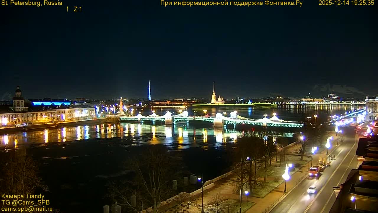 An overcast day presents a wide shot of a city along a grey river, featuring a multi-arched bridge with active traffic, a diverse cityscape including a prominent domed building and a white boat, all framed by bare trees in the foreground.