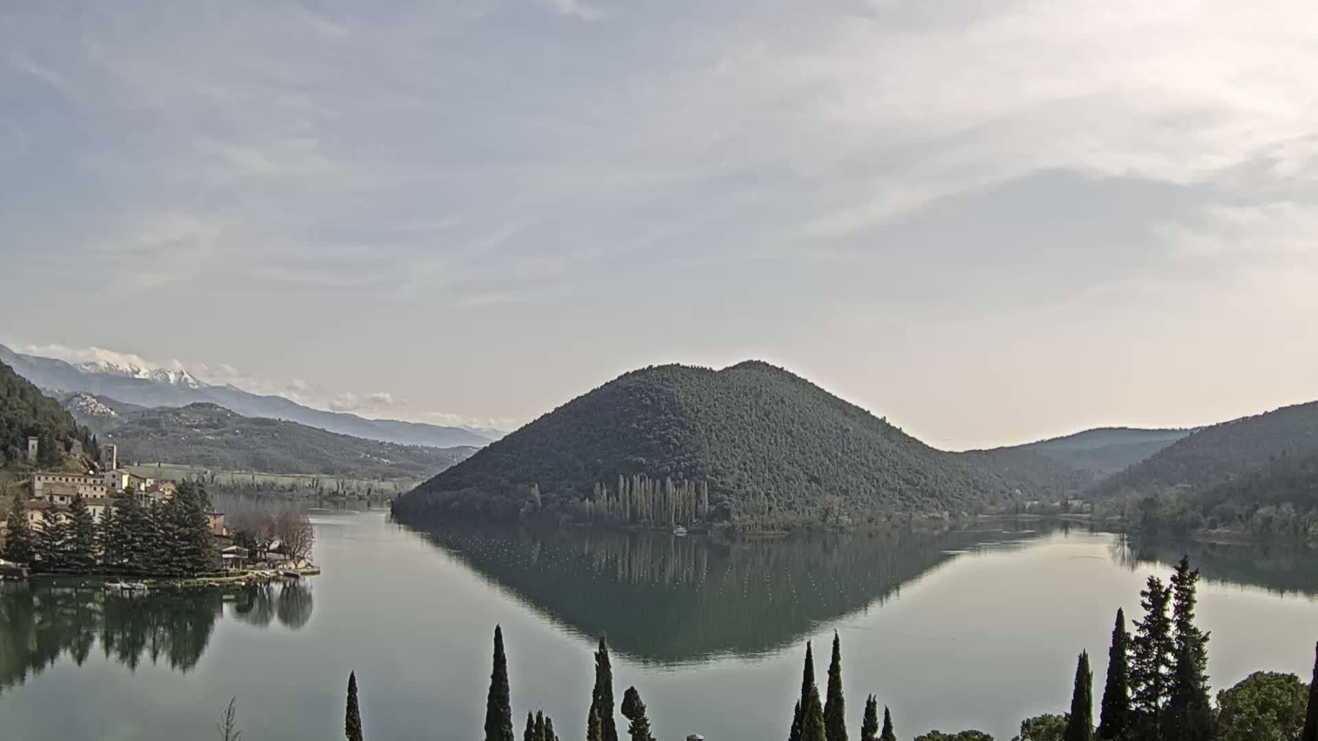 A calm lake reflects a tree-covered hill and distant snow-capped mountains under a mostly cloudy sky.