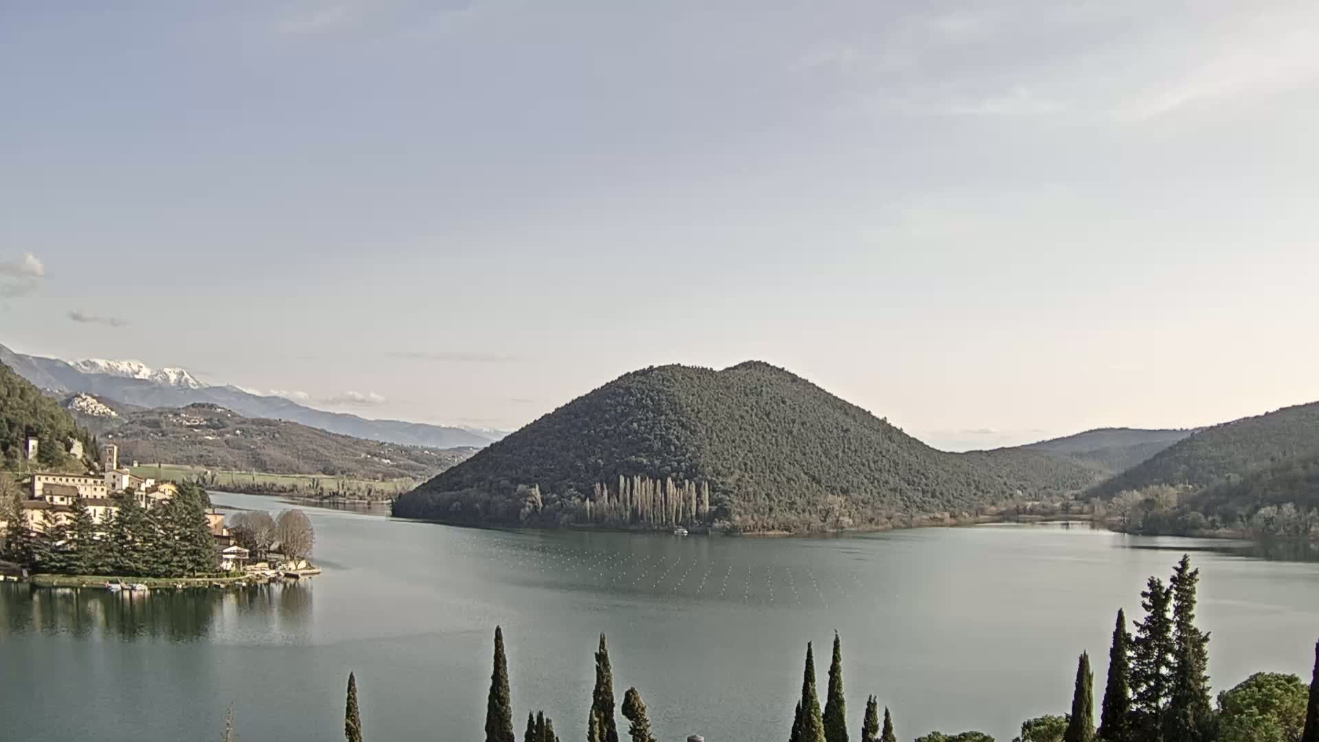 A tranquil lake, surrounded by rolling hills and mountains with some snow on the distant peaks, is under a clear, bright sky.