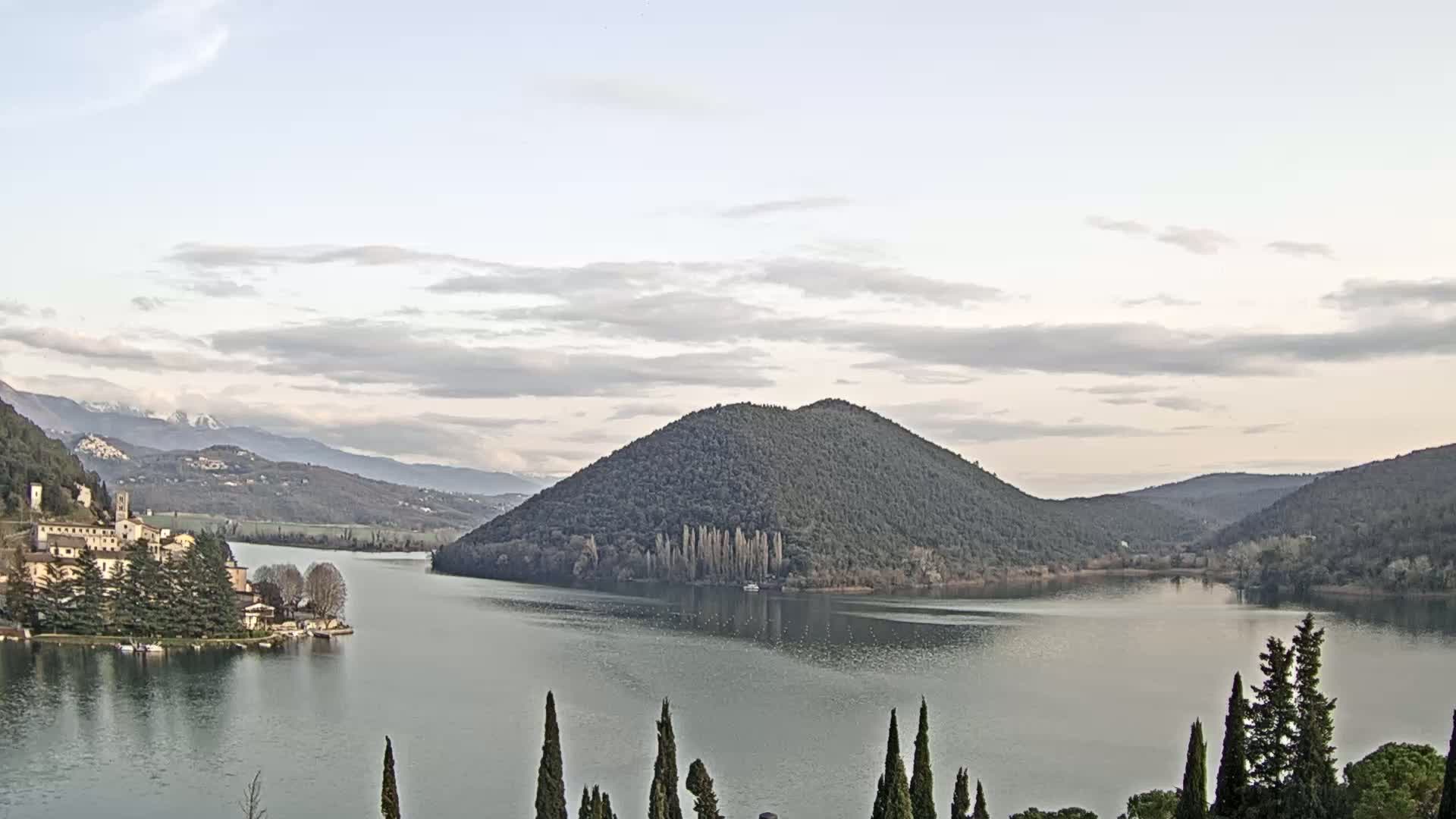 A calm lake is surrounded by hills and mountains under a partly cloudy sky, with a village nestled on the lakeshore.