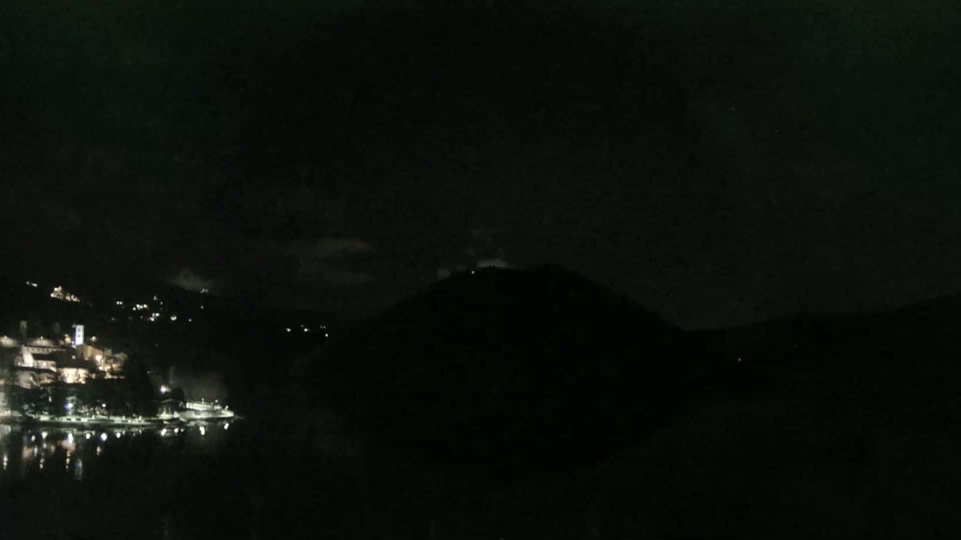 A nighttime view shows a dark hill across a body of water from a dimly lit town.