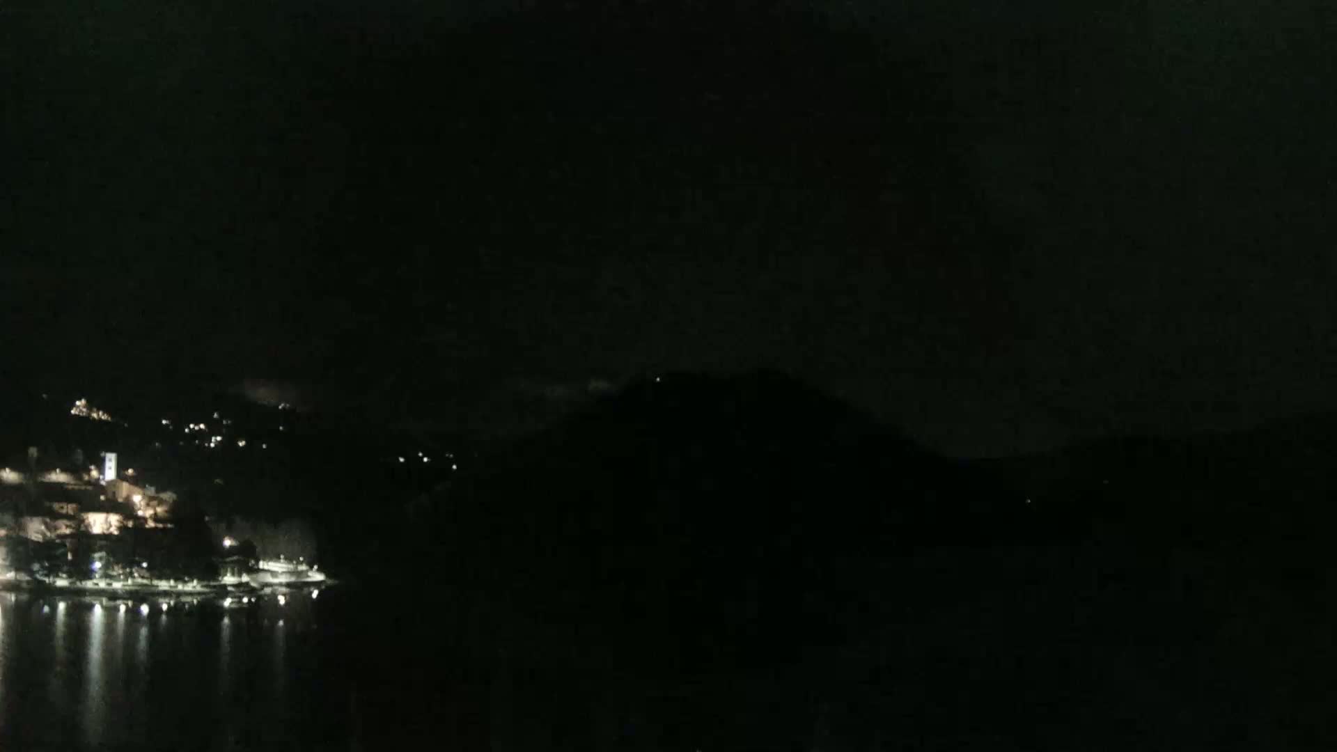 A nighttime view shows a dark, hilly landscape with a shoreline of lights reflecting in calm water under a mostly clear, dark sky.