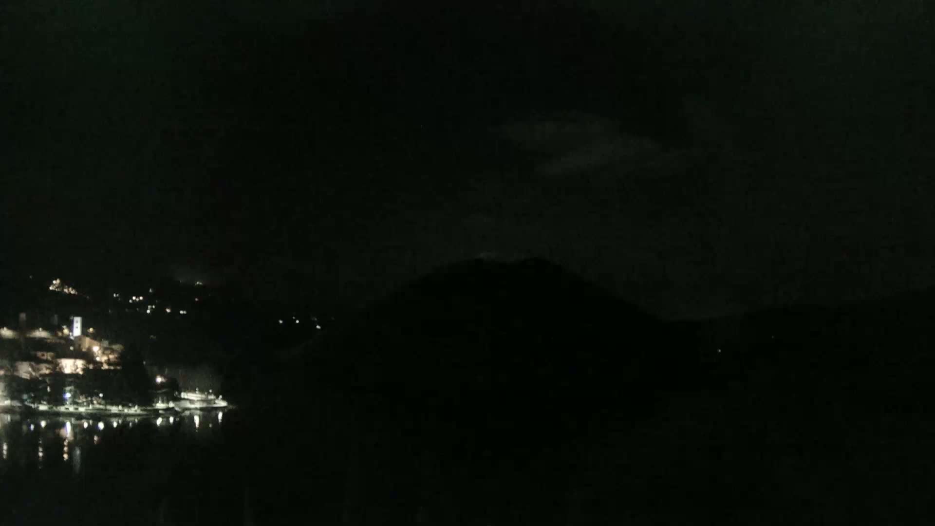 A nighttime view of a dimly lit town situated along a dark body of water, partially obscured by a mountain under a cloudy sky.