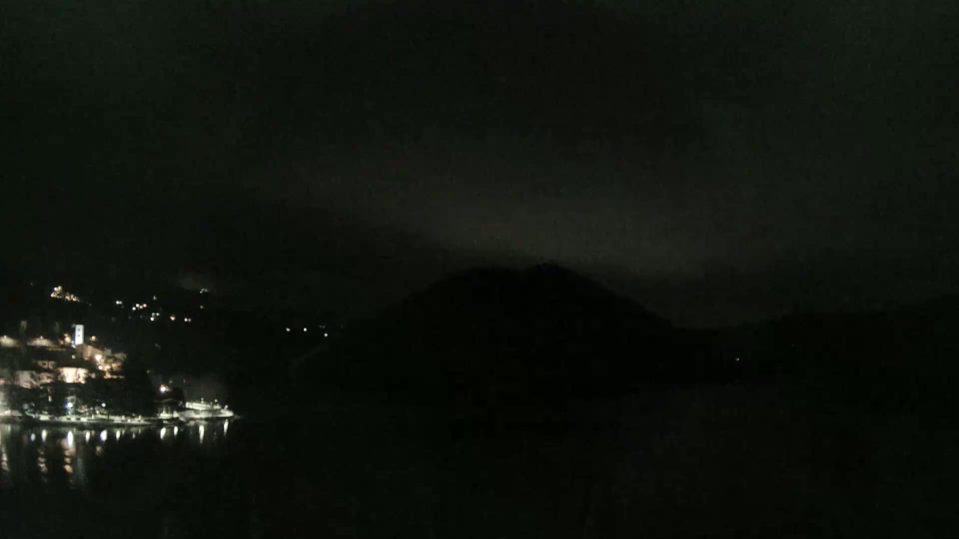 A dark, nighttime scene shows a dimly lit coastal town nestled beside a silhouetted hill, under a mostly cloudy sky.